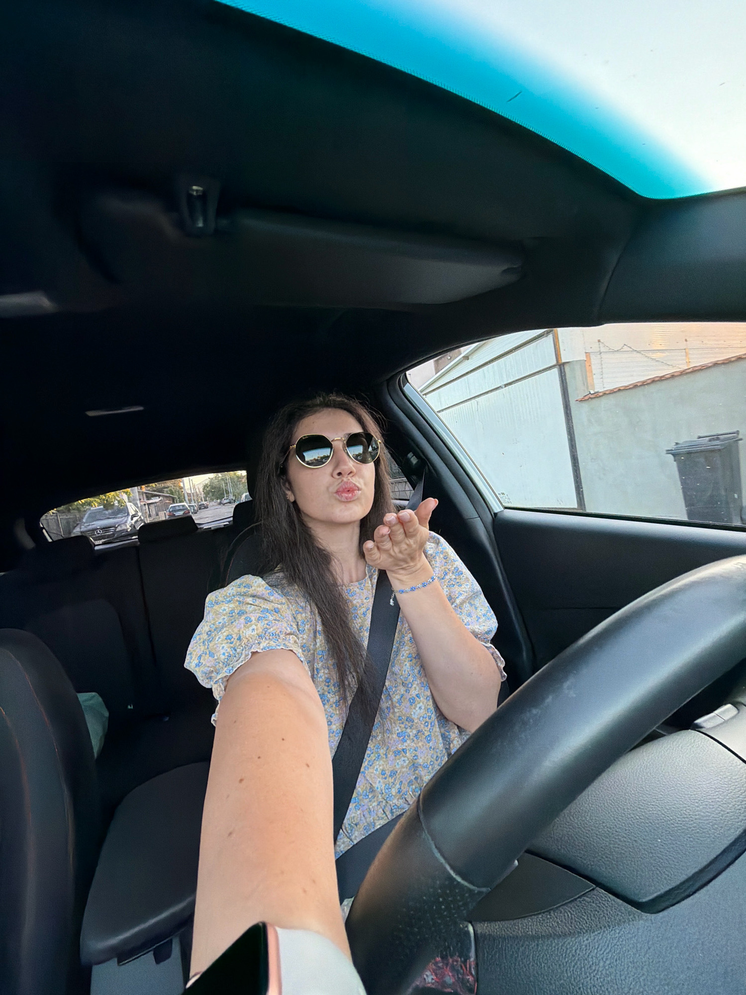 A young woman with long, dark hair is sitting in the driver's seat of a car, taking a selfie. She is wearing sunglasses and a floral print top, and she is blowing a kiss towards the camera. Her left arm is extended, holding a smartphone. The car's interior is visible, including the steering wheel, dashboard, and sunroof. Outside the car, a street scene with buildings and other cars can be seen. The image was taken in Sofia, Bulgaria.