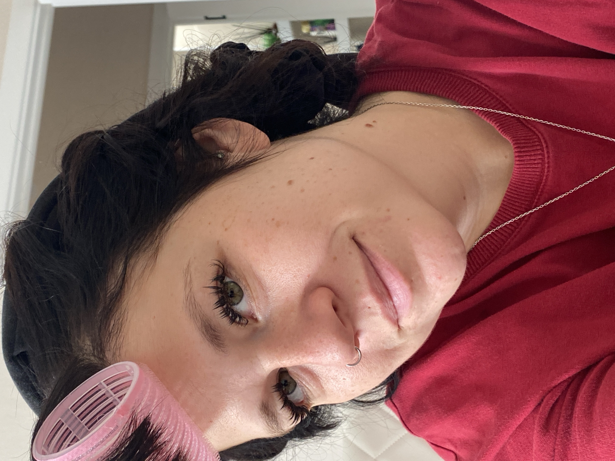 A medium shot shows a young woman with dark hair and a red shirt. She is wearing pink hair rollers in her hair and has a nose ring. She is looking towards the camera with a slight smile. The background is blurred, but appears to be an indoor setting with a doorway and shelves visible. The image quality is clear and in focus, capturing details of her facial features and attire.