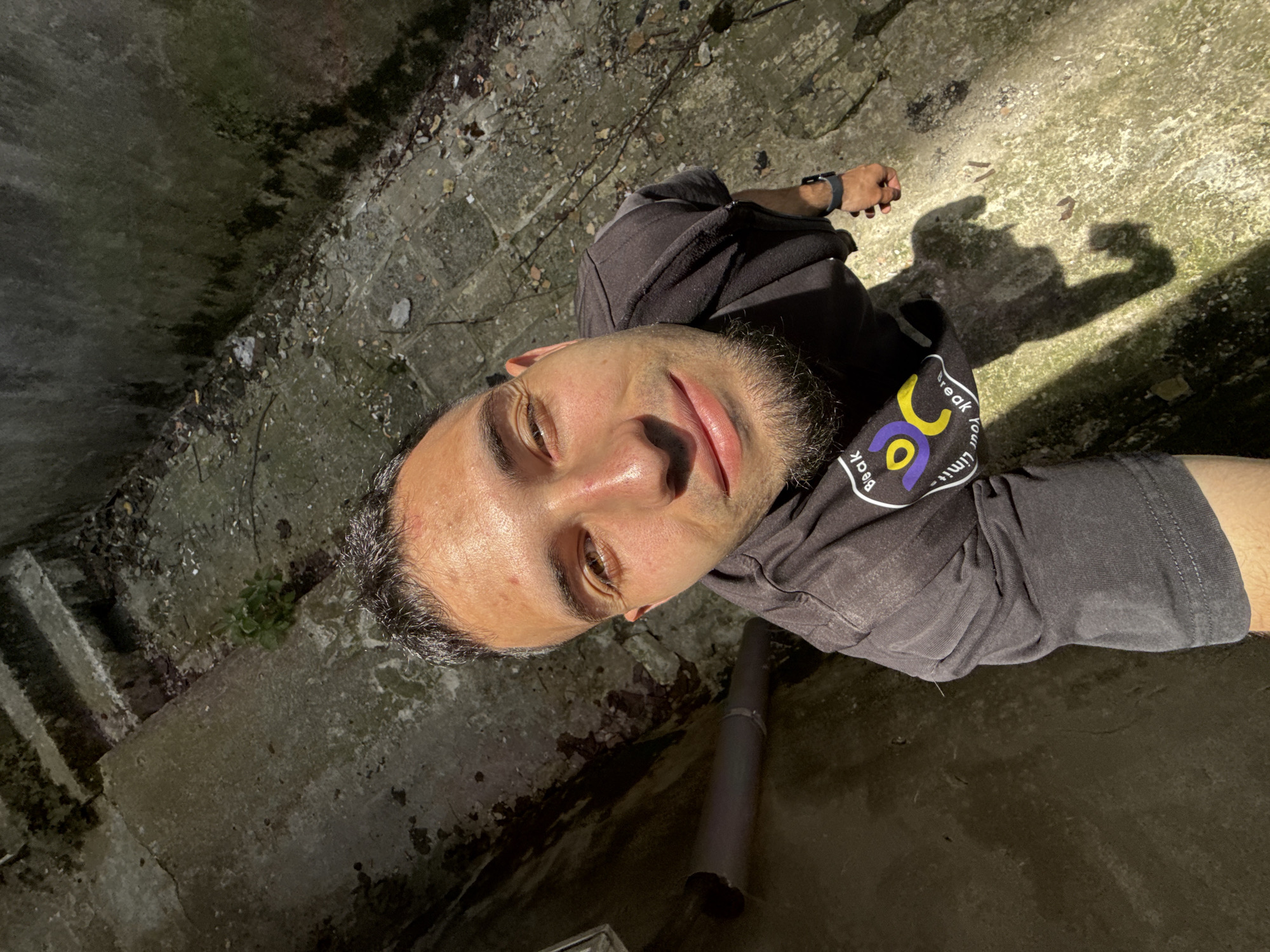A man is taking a selfie in what appears to be an abandoned or derelict urban area. The location has concrete walls and surfaces, some with visible wear and moss growth. A gray pipe lies on the ground near the man. The man is wearing a gray t-shirt and a black hoodie. He is also wearing a watch on his left wrist. The light source is casting shadows on the ground behind him. It cannot be determined from the image the location is in Shumen, Bulgaria.