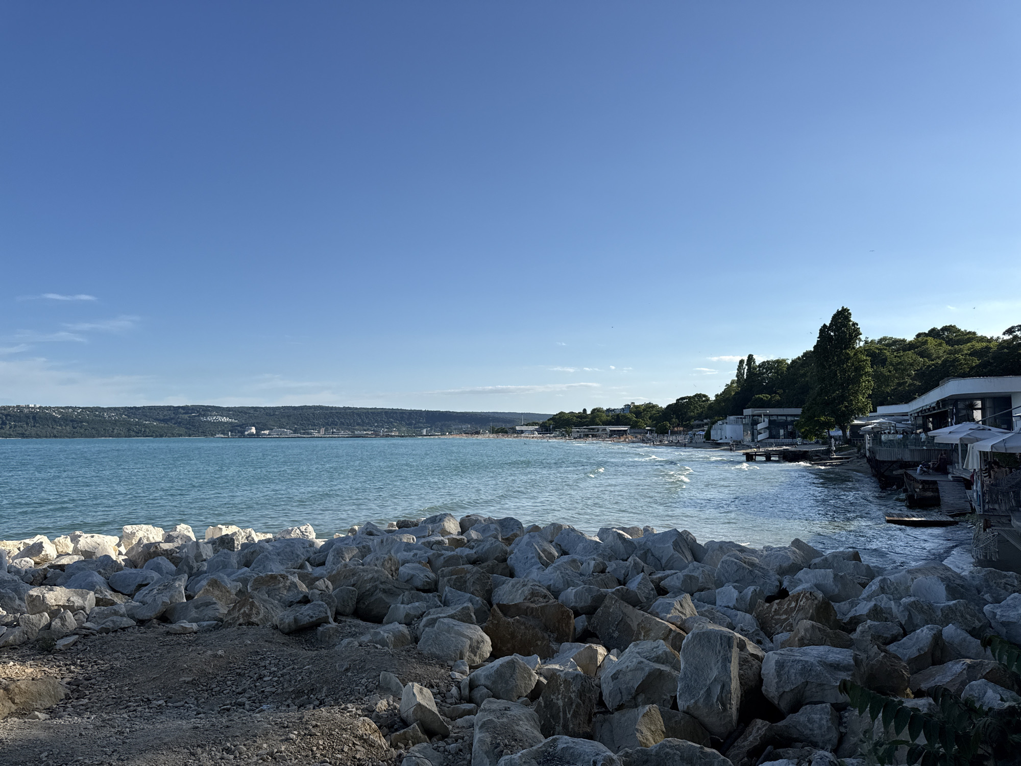 This image captures a scenic view of the Varna Bay in Varna, Bulgaria. In the foreground, a densely packed arrangement of large, irregular grey and white rocks lines the shore, with smaller gravel and dirt visible at the very bottom. The sea, a vibrant turquoise, stretches out towards the horizon, displaying gentle waves that reach the rocky embankment. Across the bay, a long stretch of sandy beach is dotted with numerous beach umbrellas and a few scattered buildings. Further in the distance, a verdant, hilly landscape forms the backdrop, with buildings and trees visible on its slopes. Along the coastline, a promenade with several structures, including outdoor seating areas with umbrellas, is situated just beyond the beach. The sky above is a clear, bright blue with a few faint wisps of clouds visible on the far left. The overall impression is one of a sunny day at a coastal resort.