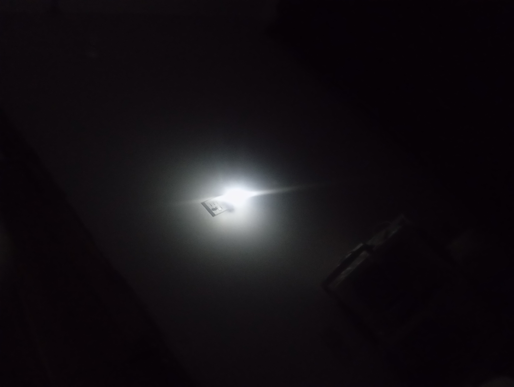 The image is a dark, low-light photograph taken from a low angle, looking upwards. A bright, square-shaped light source, possibly an LED, is visible in the upper-middle portion of the frame, emitting a strong glare that illuminates the surrounding area with a diffused light. To the right of the light source, a rectangular object with a textured surface, appearing dark and possibly metallic, is partially visible. The rest of the scene is cast in deep shadow, with indistinct shapes and a hazy atmosphere, suggesting the photograph might have been taken indoors or in an environment with poor ambient light.