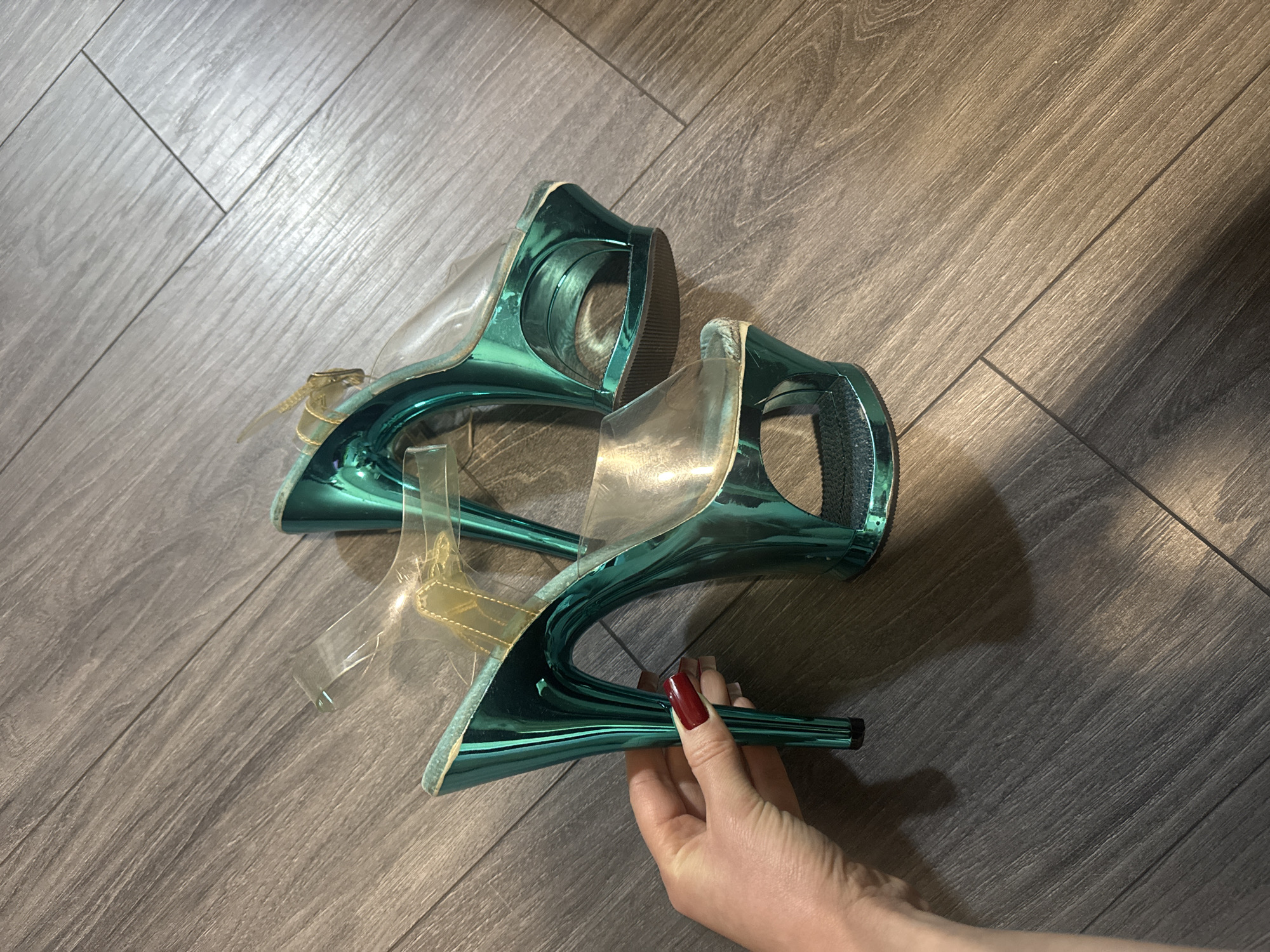 The scene is an indoor setting within Unknown, Unknown, captured from a high-angle perspective. Two platform high-heel shoes with a metallic teal-green finish are resting on dark grey-brown laminate or hardwood flooring arranged in diagonal planks. A left human hand, exhibiting light skin and long, dark red manicured fingernails, grasps the stiletto heel of the foreground shoe. Each shoe features transparent PVC toe straps and a transparent ankle strap, with a visible gold-colored buckle on the rear shoe's strap. The shoes possess tall stiletto heels and thick platforms, all coated in the metallic teal-green material. Noted wear along the edges of both platforms, with the foreground shoe's inner platform edge showing significant abrasion, exposing a lighter substrate.