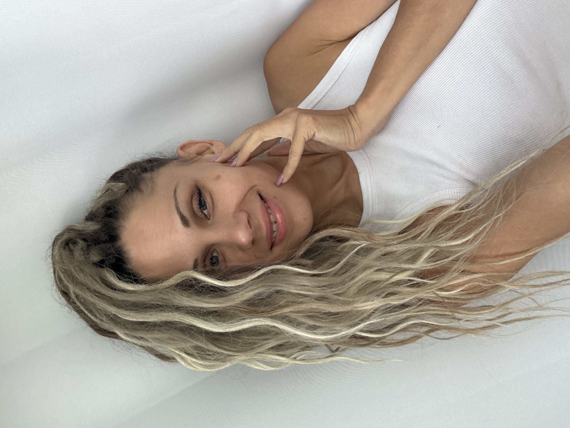 A woman with long, blonde dreadlocks styled in a high ponytail rests her hand on her cheek, smiling at the camera. She is wearing a white ribbed tank top. The background is a plain white wall, slightly creased, giving the impression of a curtain or backdrop. There is no indication of the city of Rusanovo, Russia, in the image.