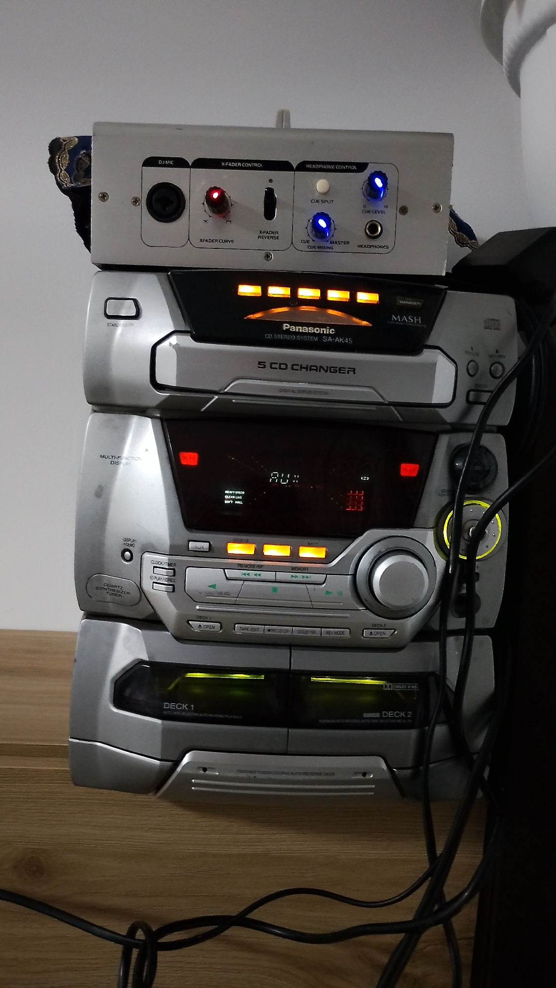 The image shows a stack of audio equipment on a wooden surface. The topmost device is a silver mixer with various controls and inputs, including a large circular input labeled "DJ-MIC". Below the mixer is a Panasonic CD stereo system with a five-CD changer and a display showing "AUX". Further down is another component of the stereo system with buttons and a large volume knob. The bottommost components are two tape decks, labeled "DECK 1" and "DECK 2", both with open bays. The equipment is silver and black, with illuminated buttons and displays. There are also black cables connected to the devices. The background is a plain white wall. The scene does not contain any people. The location of this image is not specified in the image data, therefore it cannot be determined to be in Smolyan, Bulgaria.
