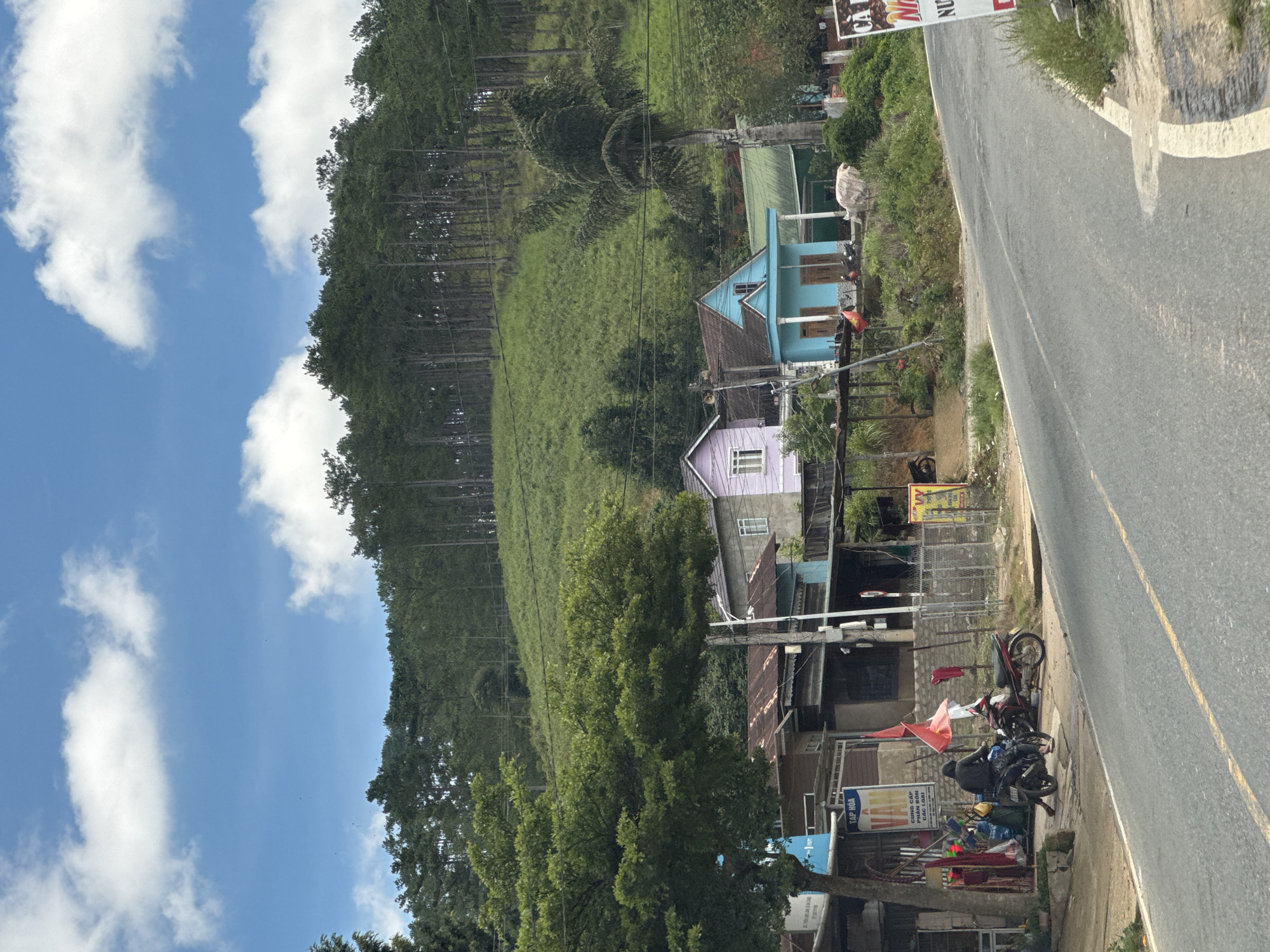 The image depicts a rural scene in Đưng K'Si, Vietnam, with a sloping, tree-covered hillside dominating the background under a blue sky with scattered white clouds. In the mid-ground, a paved road curves to the right, with a narrow sidewalk bordering it. Along the sidewalk, a series of buildings are visible.
On the left, a person in a dark hooded jacket is seated on a black motorcycle. To the right of the motorcycle, a red scooter is parked. A weathered building with a corrugated metal roof is situated behind the motorcycles, with various items displayed outside, including what appears to be clothing and bags. A sign with Vietnamese text is also visible on this building.
Further to the right, two distinct houses stand out: one is painted lavender with white trim and windows, and the other is a bright turquoise with white trim and pillars. Both houses have pitched roofs. Behind these houses, more buildings with green roofs and various trees and vegetation are present. A tall palm tree stands to the right of the turquoise house.
In the far right of the image, a portion of a large white sign with red text is visible, advertising "CA NHUNG" and "NU". The overall impression is one of a quiet, somewhat rustic settlement nestled at the base of a forested hill.