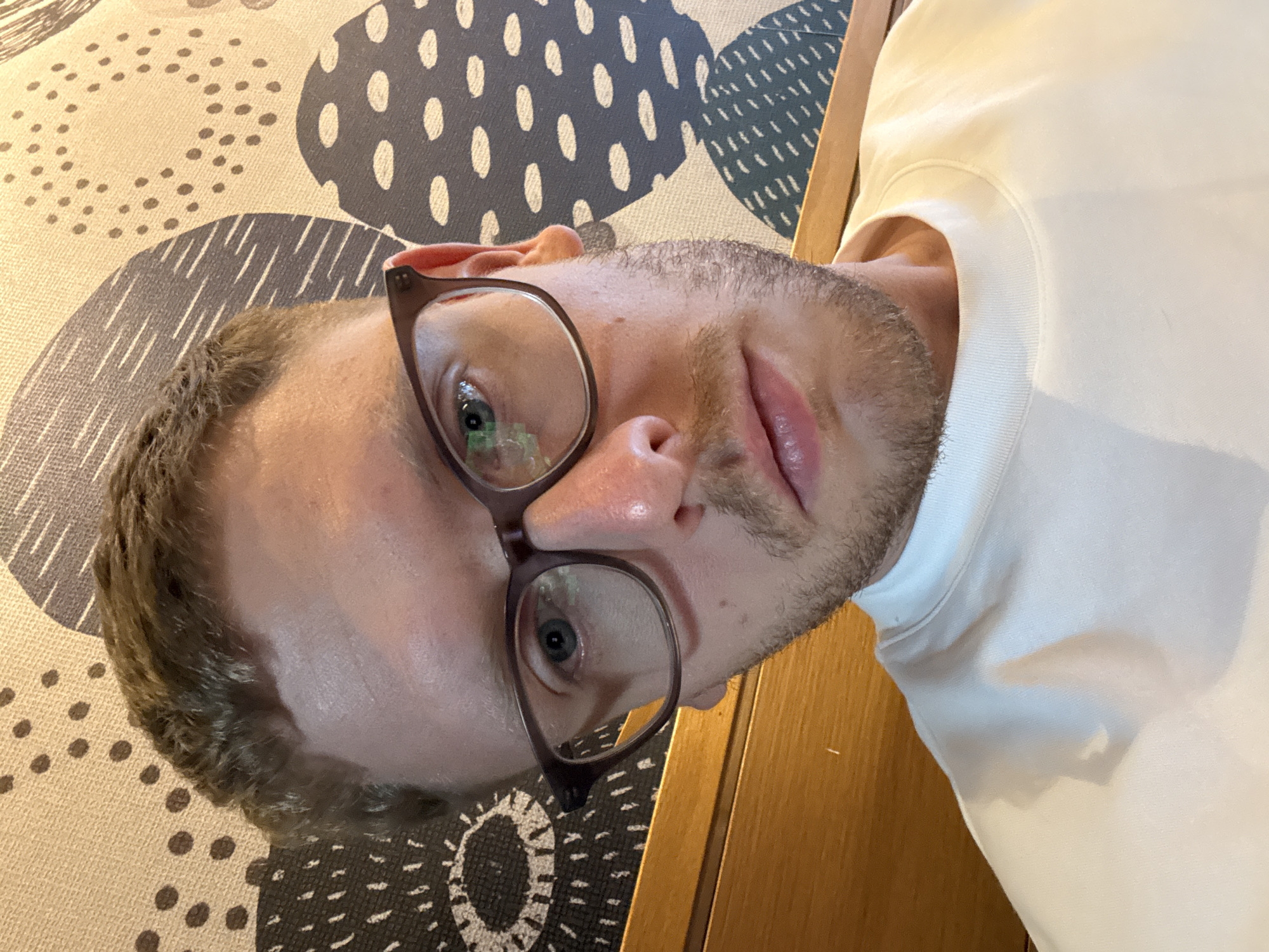 A rotated selfie shows a man with fair skin, light brown hair, and a short beard. He is wearing glasses with a dark brown frame and a white sweatshirt. The background includes a patterned rug or mat with circular and lined designs in shades of beige and gray. A wooden surface is visible in the lower right corner. The location in the city of Beşiktaş, Turkey, cannot be determined from the visual information provided.