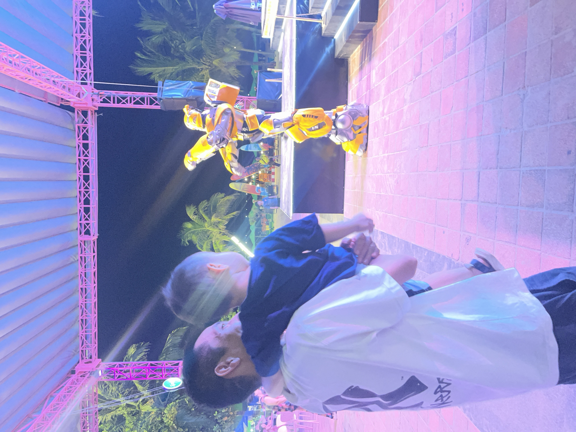 In this night scene, a person carries a young child, who is looking towards a costumed performer dressed as Bumblebee, a character from the Transformers franchise. The Bumblebee performer stands on a stage, striking a dynamic pose with one leg extended. The setting appears to be an outdoor entertainment venue, possibly along the coast in Da Nang, Vietnam, indicated by the presence of palm trees and a bright, artificial lighting scheme that bathes the scene in pink and blue hues. The ground is paved with square tiles. In the background, there are structures that suggest a stage setup and possibly other attractions or amenities associated with a tourist area.
