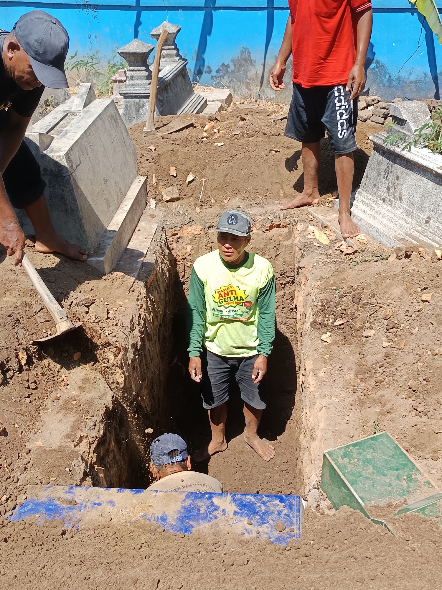 In the city of Geneng, Indonesia, a scene unfolds in what appears to be a cemetery. Four individuals are present, engaged in activities related to a grave. One person, wearing a gray cap and a yellow long-sleeved shirt with black shorts, stands in a freshly dug trench. Another individual, also wearing a gray cap and a light-colored shirt with dark shorts, is positioned inside the trench, partially obscured by a blue tarp. A third person, clad in a red t-shirt and black Adidas shorts, stands on the edge of the grave. The fourth person, visible from the chest up and wearing a dark shirt and a gray baseball cap, is bent over, working with a shovel near a concrete tombstone. The environment is characterized by mounds of soil, exposed earth from the excavation, and several concrete grave markers. The background features a bright blue wall and more grave structures, indicating an outdoor, likely sunny, setting.