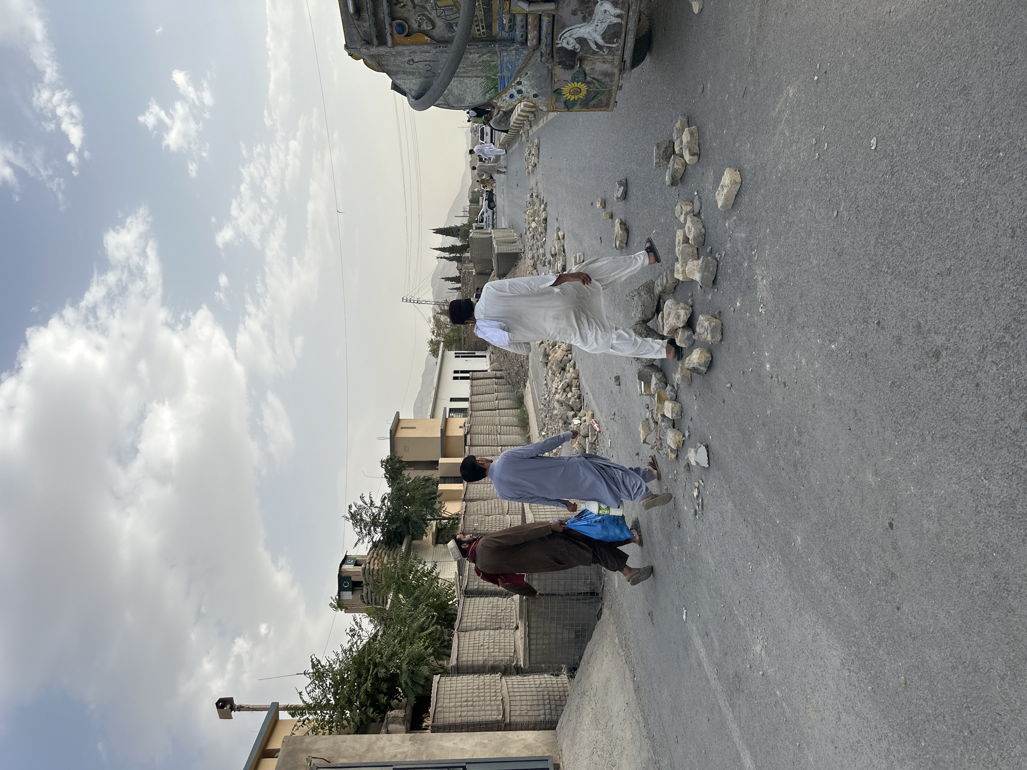 Three men walk along a paved road in Quetta City Tehsil, Pakistan. The man on the left is wearing a brown robe and a red scarf. The man in the middle is wearing a light blue robe and carrying two blue plastic bags. The man on the right is wearing a white robe and has a white cloth draped over his left shoulder. Bricks are scattered across the road in front of the men. To the left of the road, stacked sandbags form a barrier, and behind them, buildings and trees are visible. On the right side of the road, a colorful, decorated truck is parked, and more people are visible further down the road. The sky is cloudy.