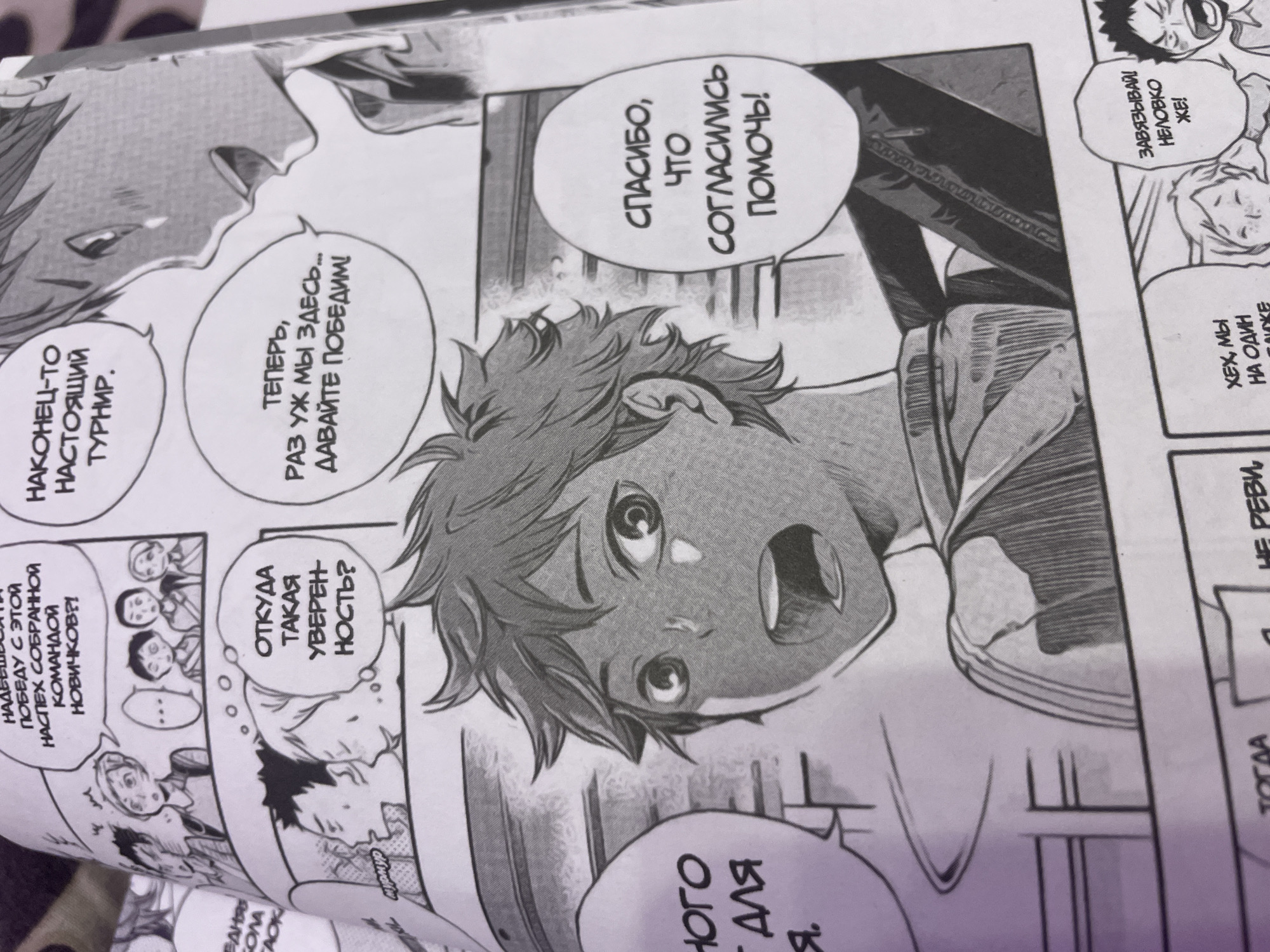 The image is a panel from a manga, likely depicting a scene from the anime/manga series Haikyuu!!, given the art style and character designs. It shows several characters in what appears to be a sports context, likely volleyball.
In the foreground, a young male character with spiky orange hair and wide, expressive eyes is depicted from the chest up, looking surprised or determined. He has a slightly flushed face.
Above and to the left of him, a speech bubble contains Russian text: "ОТКУДА ТАКАЯ УВЕРЕННОСТЬ?" which translates to "Where does such confidence come from?".
To the right and slightly above this, another speech bubble contains Russian text: "НАКОНЕЦ-ТО НАСТОЯЩИЙ ТУРНИР." which translates to "Finally, a real tournament.".
Further to the right, another speech bubble, directed towards the orange-haired character, has Russian text: "СПАСИБО, ЧТО СОГЛАСИЛИСЬ ПОМОЧЬ!" which translates to "Thank you for agreeing to help!".
In the background, there are other characters depicted in smaller panels and silhouettes, suggesting a team or competition setting. One group of characters is shown looking at the orange-haired character with varying expressions.
The text within the manga is in Russian.
The image quality suggests it is a photograph of a printed manga page, possibly from a Russian edition. There are no visible elements that tie this scene to the city of Irinovka, Russia, or any specific urban environment. The setting appears to be an indoor sports facility or a similar location implied by the context of the manga.