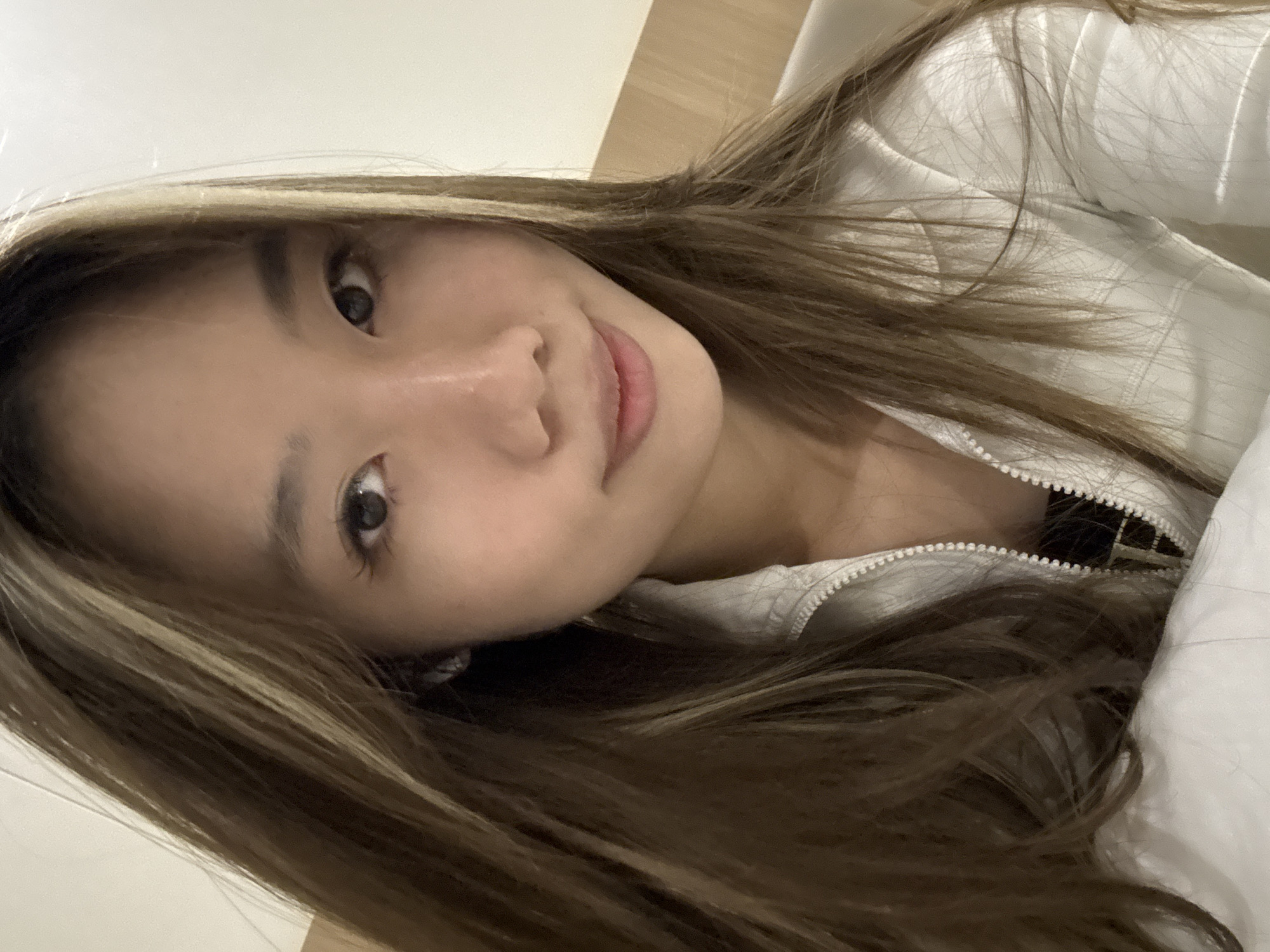 A close-up, slightly angled portrait captures an Asian woman with long, brown hair highlighted with blonde streaks. She has fair skin, brown eyes, and a slight smile. She appears to be wearing a white zip-up jacket. The background is blurred and features a light-colored wall and a white surface, possibly a bed. The image gives no indication of the location of Dunhuang City, China.