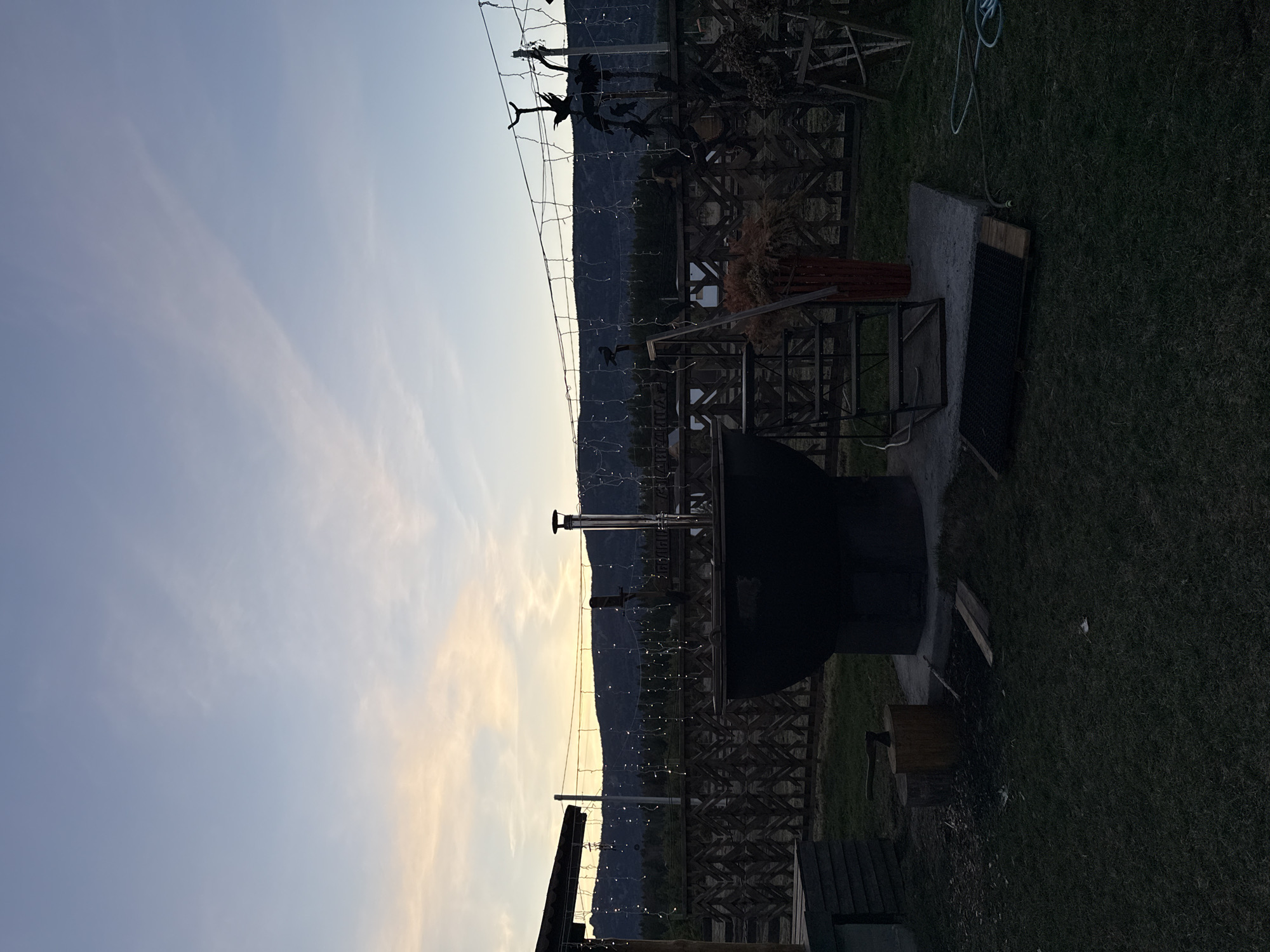 The image shows an outdoor scene with a metal structure and what appears to be a wood-burning hot tub or stove. The background features a mountain range and a sky with some clouds. The scene is possibly located in Tyulyuk, Russia, based on the prompt. There's a grassy area with some wooden elements nearby the metal structure. The lighting suggests it could be dusk or dawn.