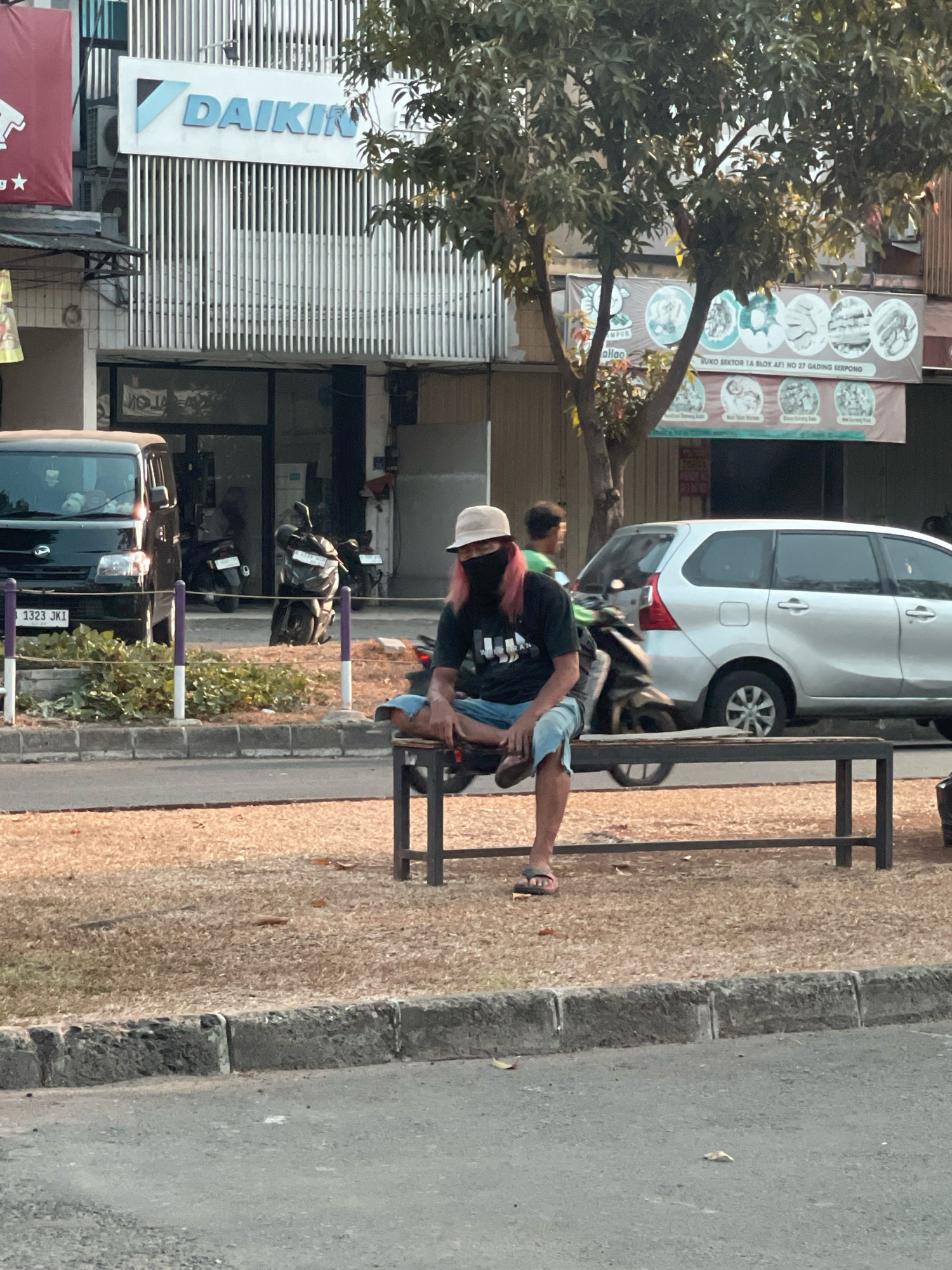 An individual with pink hair, wearing a beige hat, a dark t-shirt, and light-colored shorts, sits on a metal bench in what appears to be a public waiting area or bus stop in front of commercial buildings. The person is wearing a dark face mask and flip-flops. Behind the individual, several motorcycles are parked, and a black van is visible on the left. A silver minivan is parked on the right side of the road. The background shows the facade of a building with the "DAIKIN" logo, along with banners displaying food items. The scene is set along a street in Pakulonan Barat, Indonesia, with dry grass and a concrete curb bordering the road.