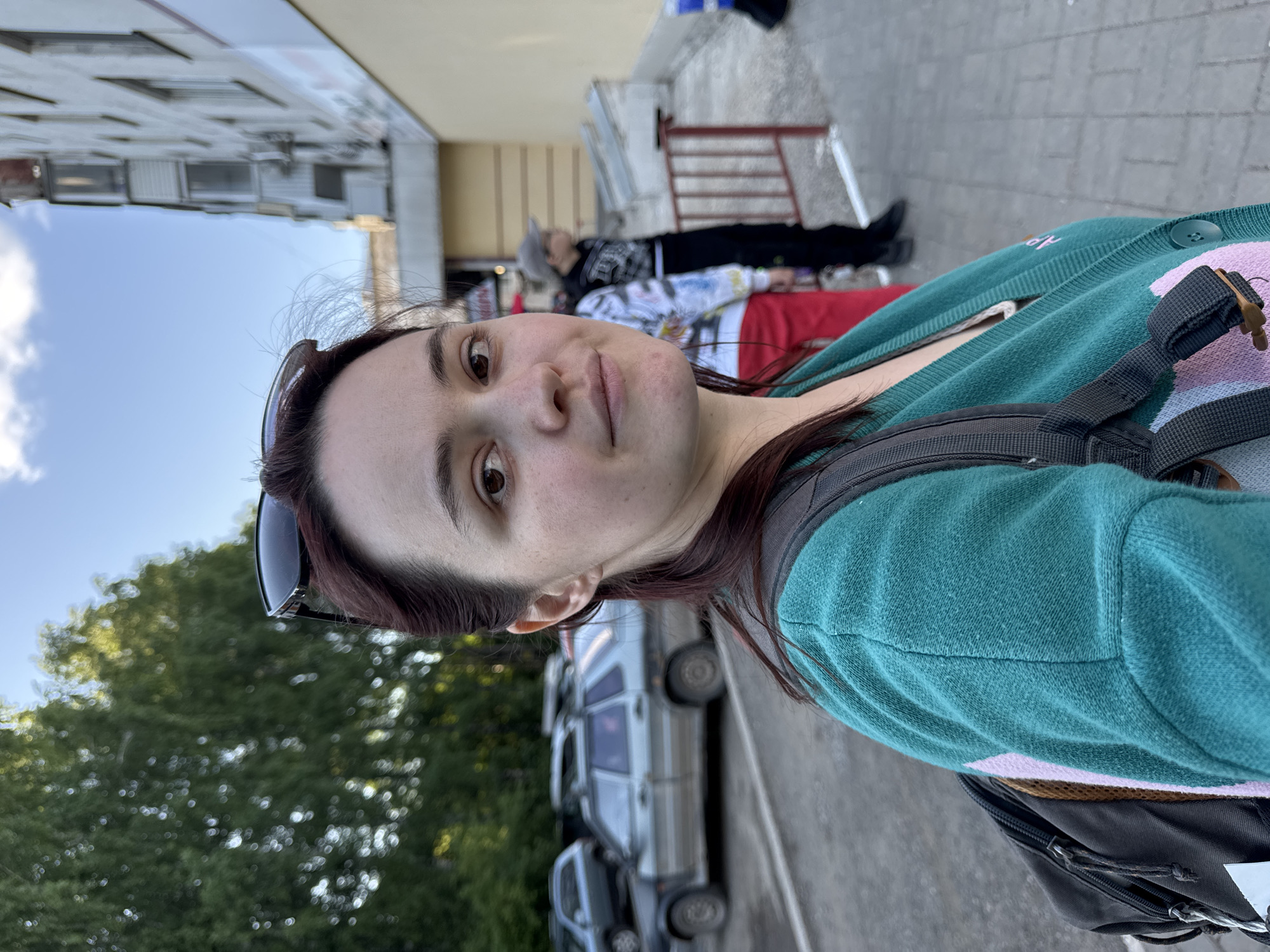 A young woman with dark brown hair and fair skin is prominently featured in a close-up, angled shot, looking directly at the viewer with a calm expression. She wears sunglasses pushed up onto her head and a teal or dark green cardigan with subtle pink or light purple patterns on the side. A black backpack is visible over her shoulder.
The setting appears to be an urban street in Izhevsk, Russia, during daytime under a clear sky with scattered clouds. In the background, two other individuals are visible further down the street. One person wears a light-colored beanie and a patterned jacket, while another is dressed in red pants. They appear to be standing or resting near a building with light-colored walls and some red metal railings.
Parked cars, possibly vans, line the side of the road behind the main subject, and green trees are visible in the distance, indicating an outdoor environment. The lighting suggests it is bright, likely morning or afternoon. No legible text is visible on any signs, clothing, or objects within the image.
