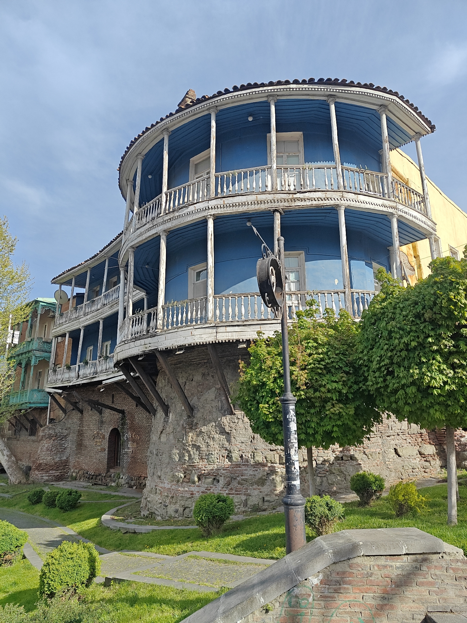 The image displays a picturesque scene in what appears to be a historic quarter, characterized by traditional architecture and lush greenery. The central subject is a multi-story building featuring distinctive blue walls and elaborate white wooden balconies that curve gracefully, encompassing a semi-circular or circular facade. The balconies are supported by slender columns and adorned with ornate balustrades and intricate carvings along their edges. The white paint on the woodwork shows some weathering, adding to its aged character.
To the left, a portion of another building with similar wooden balconies, painted in green and light brown, is visible. To the right, a section of a yellow building abuts the blue structure. The main blue building rests on a sturdy foundation of old stone and brick walls, with wooden beams extending from its base.
In the foreground and midground, a landscaped area with well-maintained green grass, small bushes, and several trees with vibrant green leaves is visible. A dark, ornate lamppost with an old-fashioned bracket stands prominently in front of the blue building. Paved pathways meander through the grassy areas, and low brick and stone walls define different sections of the garden.
The scene is captured during the daytime under a clear, bright blue sky with a few wispy clouds, suggesting sunny weather, likely in spring or summer. The lighting is soft but distinct, highlighting the architectural details.
Visible text on a small wooden sign above a door in one of the stone walls reads "Old City Wall" and partially "Restaurant" below it. The architectural style, with its characteristic wooden balconies, is indicative of old Tbilisi, Georgia.