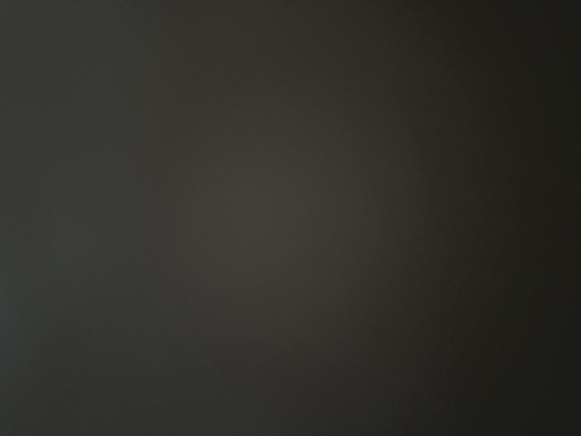 This is a photograph of a dark, textured surface, possibly a wall or fabric. The lighting is dim, with a gradient of darkness from left to right, suggesting a light source from the left side of the frame. There are no discernible subjects, objects, activities, or events taking place. The image lacks any specific details about time of day, weather, location cues, or emotions. No text is visible. The overall impression is of a minimalist, abstract image due to the absence of identifiable features.