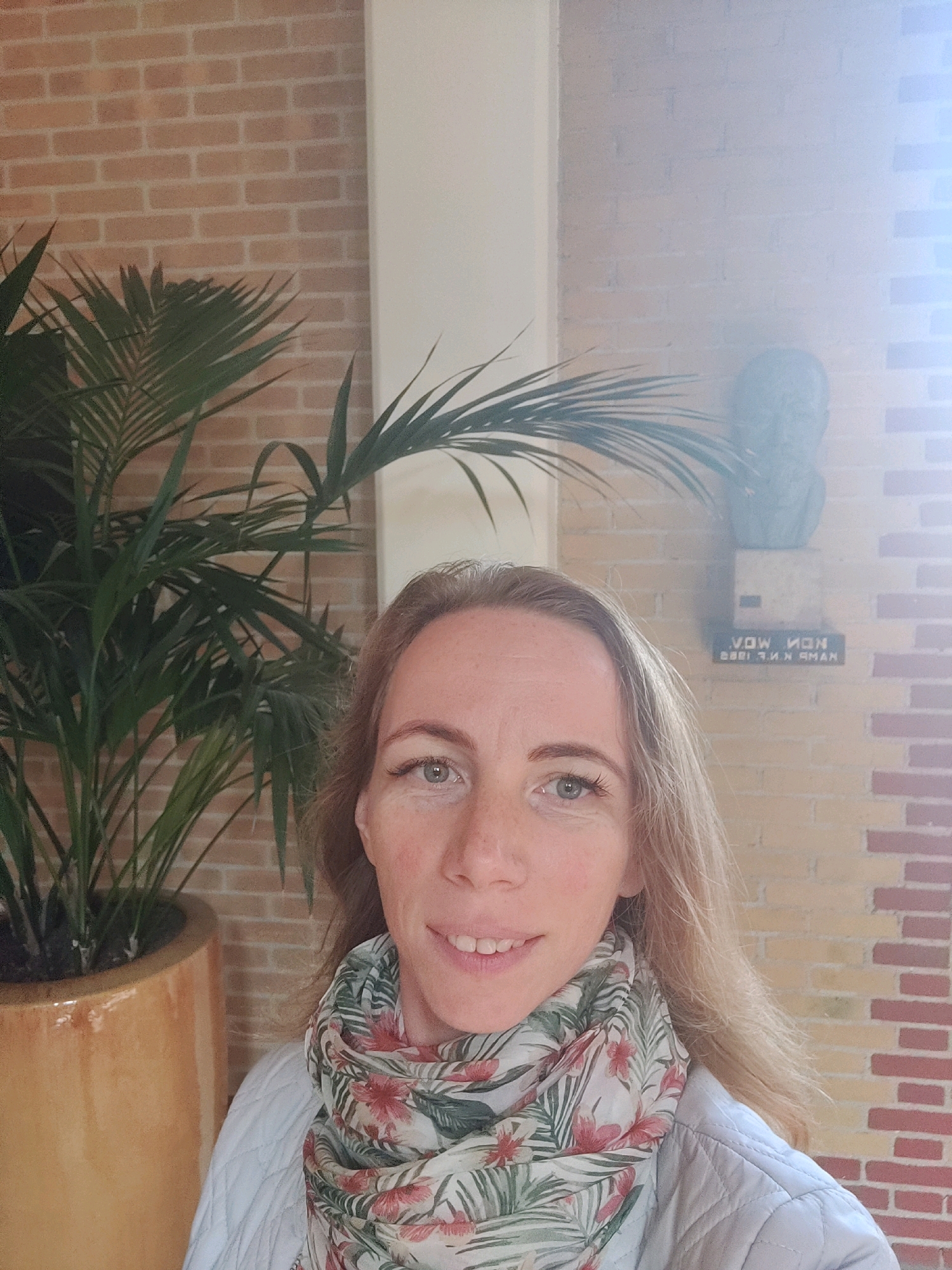A woman with blonde hair and a floral scarf smiles at the camera. She is standing in front of a brick wall and a large potted plant. A bust is mounted on the wall behind her. The text on the plaque below the bust is partially obscured but appears to read "VOW NON / 8881 F. N. K. AMAN". The location is the City Hall of Winterswijk, Netherlands.