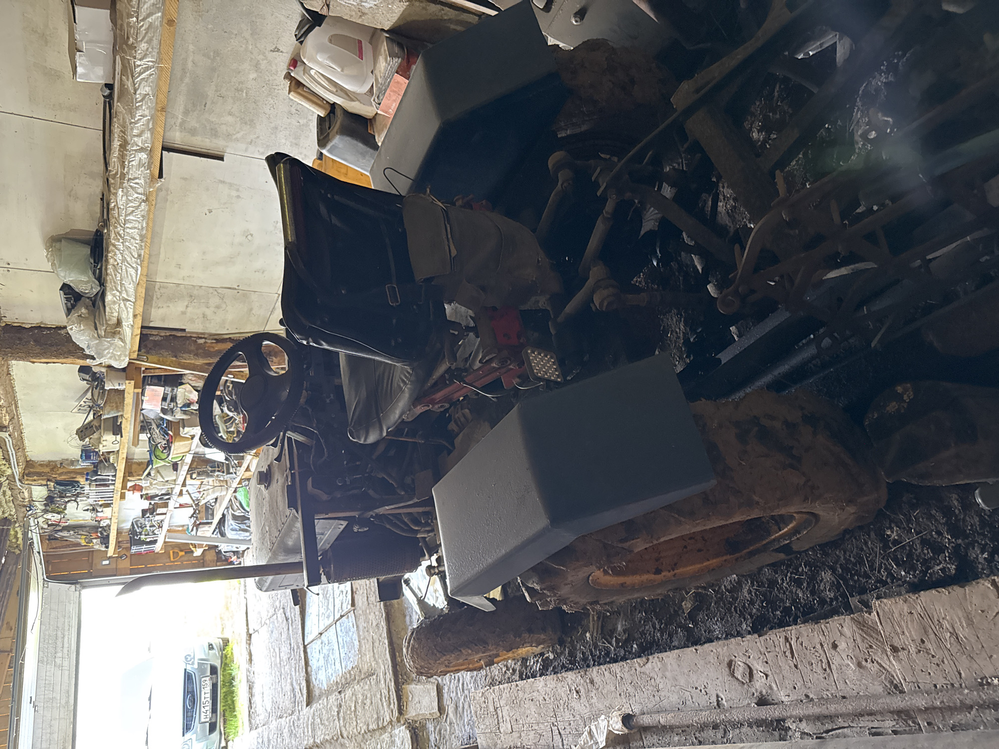 The image depicts a cluttered garage environment in Borodulino, Russia. A weathered, dark-colored mini-tractor is the central subject, positioned on a muddy floor. The tractor features large, mud-caked tires, a steering wheel, a seat, and a prominent exhaust pipe. Various items are stored on shelves in the background, including containers, tools, and other unidentifiable objects, adding to the disarray. A car, partially visible through the open garage door, is parked outside. The overall scene suggests a functional but unkempt workspace.