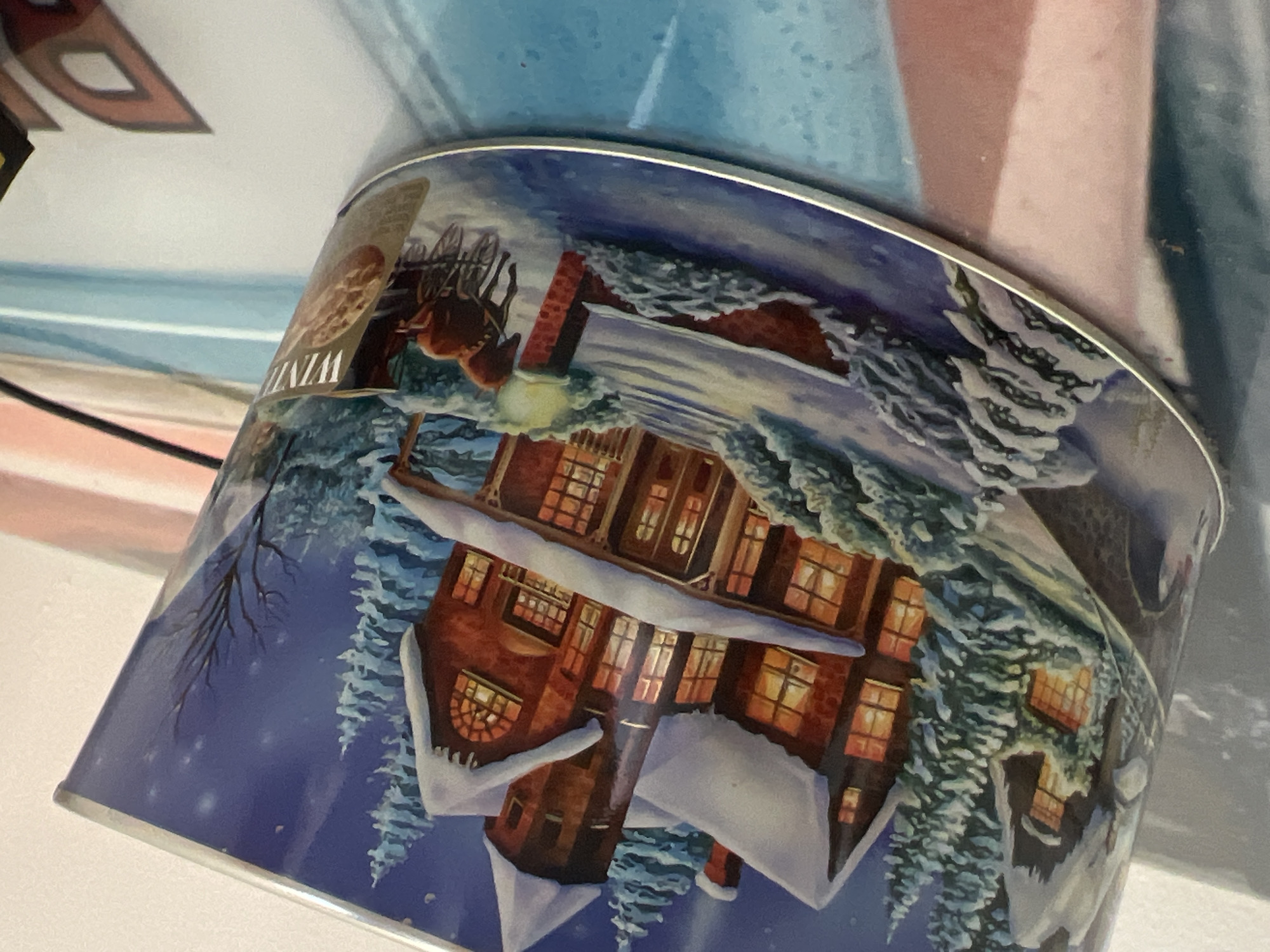 The image shows a tin decorated with a Christmas scene. The scene depicts a snow-covered house with lights on in the windows, surrounded by snow-laden evergreen trees. A horse-drawn carriage is visible in the foreground, along with a lamppost. A label on the tin partially reads "WINTER". The tin is tilted and positioned on a surface that appears to be a table or desk, with some of its reflective surface visible. The background is blurred, but shows a light-colored wall and possibly some other objects. The overall impression is of a festive, winter-themed tin, likely containing cookies or similar treats. The image does not contain any recognizable landmarks or details that would indicate its location in Balbriggan, Ireland.