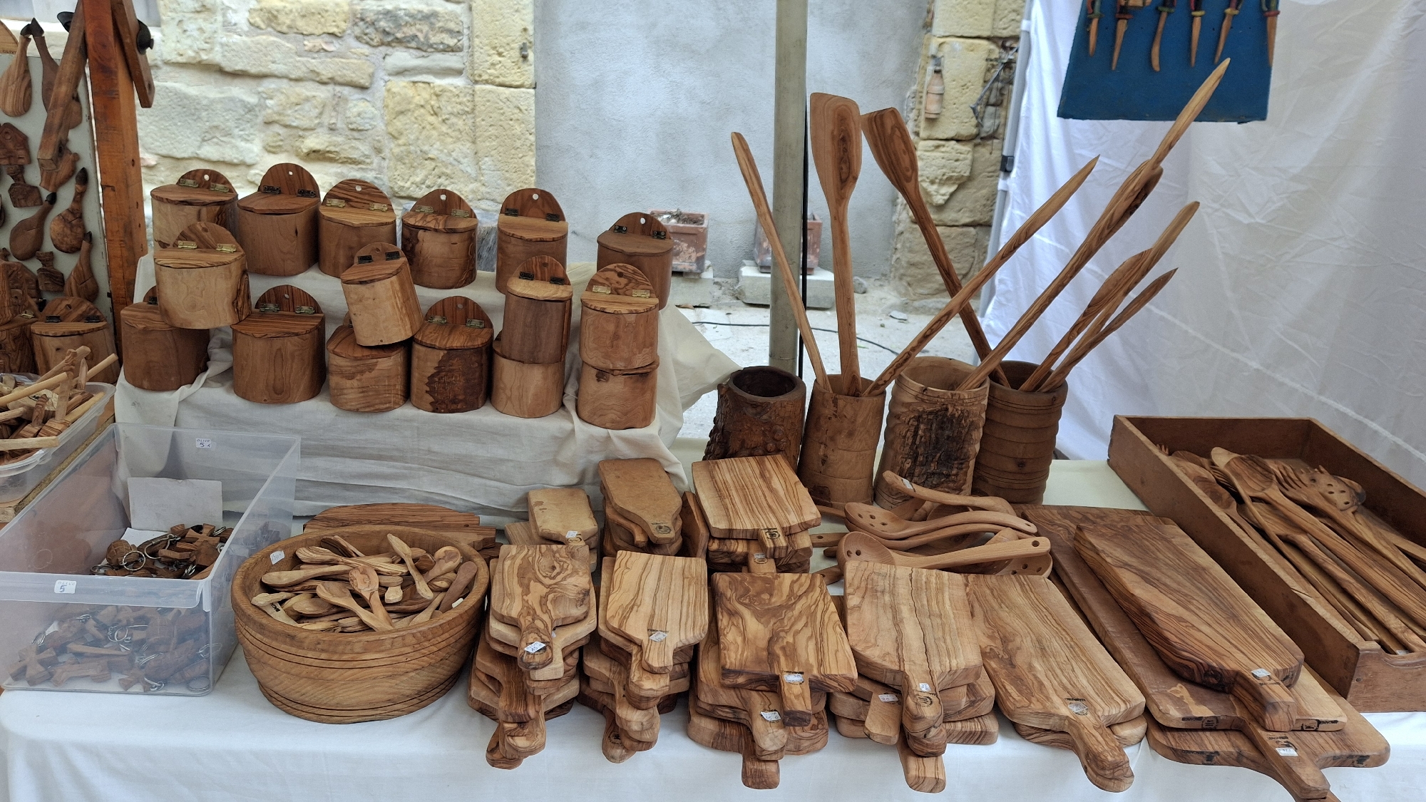 This image shows a stall at an outdoor market, likely in La Fresneda, Spain, displaying a variety of handcrafted wooden items. The stall is covered by a white canopy. In the foreground, on a white tablecloth, are stacks of small wooden cutting boards and a large wooden bowl filled with small wooden spoons. To the left, a clear plastic bin contains wooden keychains and other small wooden objects. Behind this, a display rack holds more carved wooden items. Further back, wooden pots or containers are stacked, and tall wooden utensils such as spoons and spatulas are displayed in wooden cylinders. The background shows a stone wall and a light-colored wall. There are no people visible in the image.