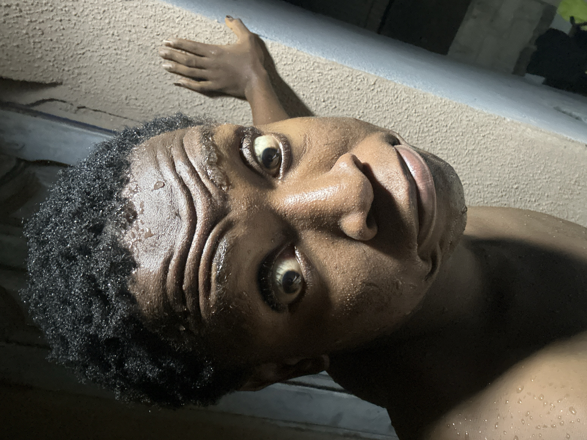 A close-up, low-angle shot of a Black person's face and partial upper body, with water droplets visible on the skin. The person is looking upwards and to the side. Their hair is short, dark, and curly. A hand is visible extended behind their head towards a grey ledge. The location and city are unknown.