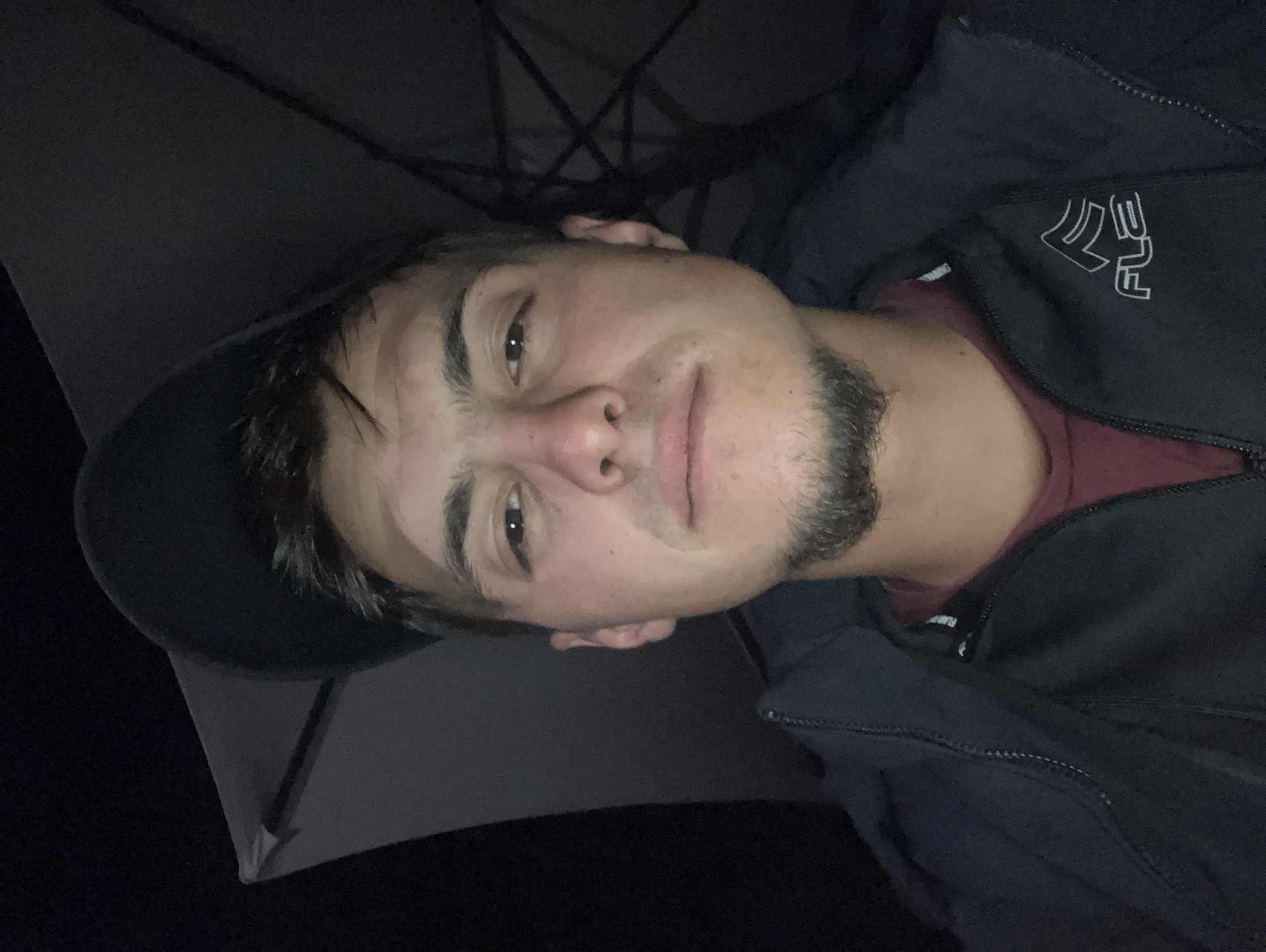 The image shows a man indoors, possibly inside a tent. He is wearing a dark-colored jacket over a maroon shirt, and a dark cap. He has brown hair, light skin, and a beard. The background of the image includes tent poles. The location is not discernible as Krastevich, Bulgaria. There are no clear actions or interactions depicted.