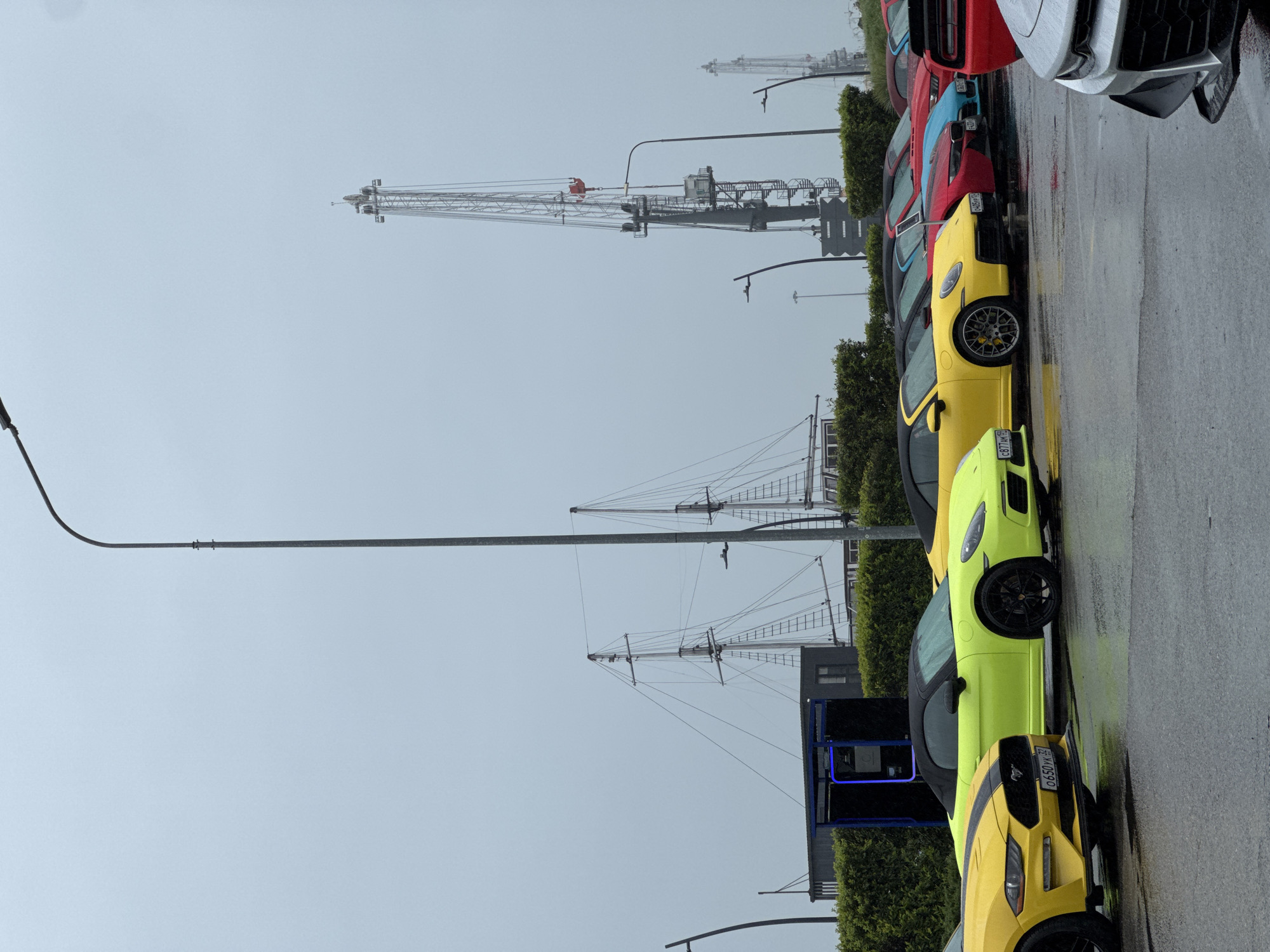 The image captures a scene on a wet, overcast day. A row of parked cars, predominantly high-end sports cars, lines a street next to a waterfront area. The cars are various colors, including yellow, green, red, blue, and silver. The street surface is wet, reflecting the overcast sky. In the background, two tall sailing ships are visible, and a large crane stands prominently in the distance, suggesting industrial port activity. The presence of the sailing ships suggests a maritime-themed area or museum. There are trees between the ships and the street. A small blue box is drawn around the back part of a bus parked in front of the ships.