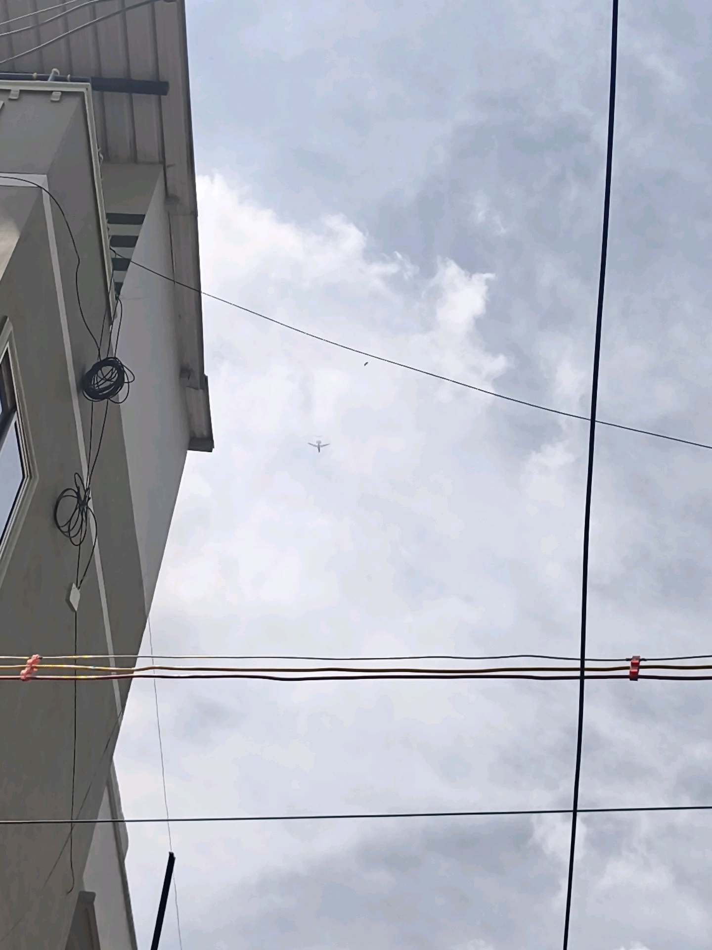 The image captures a cloudy sky over Coimbatore, India, with a residential building on the left and several electrical wires crisscrossing the frame. A small airplane is visible in the distance, flying high in the sky. The building appears to be a multi-story structure with gray siding and a metal roof. Electrical wires are bundled and attached to the side of the building, with some strung across the view. The overall scene is overcast and appears to be during daylight hours.