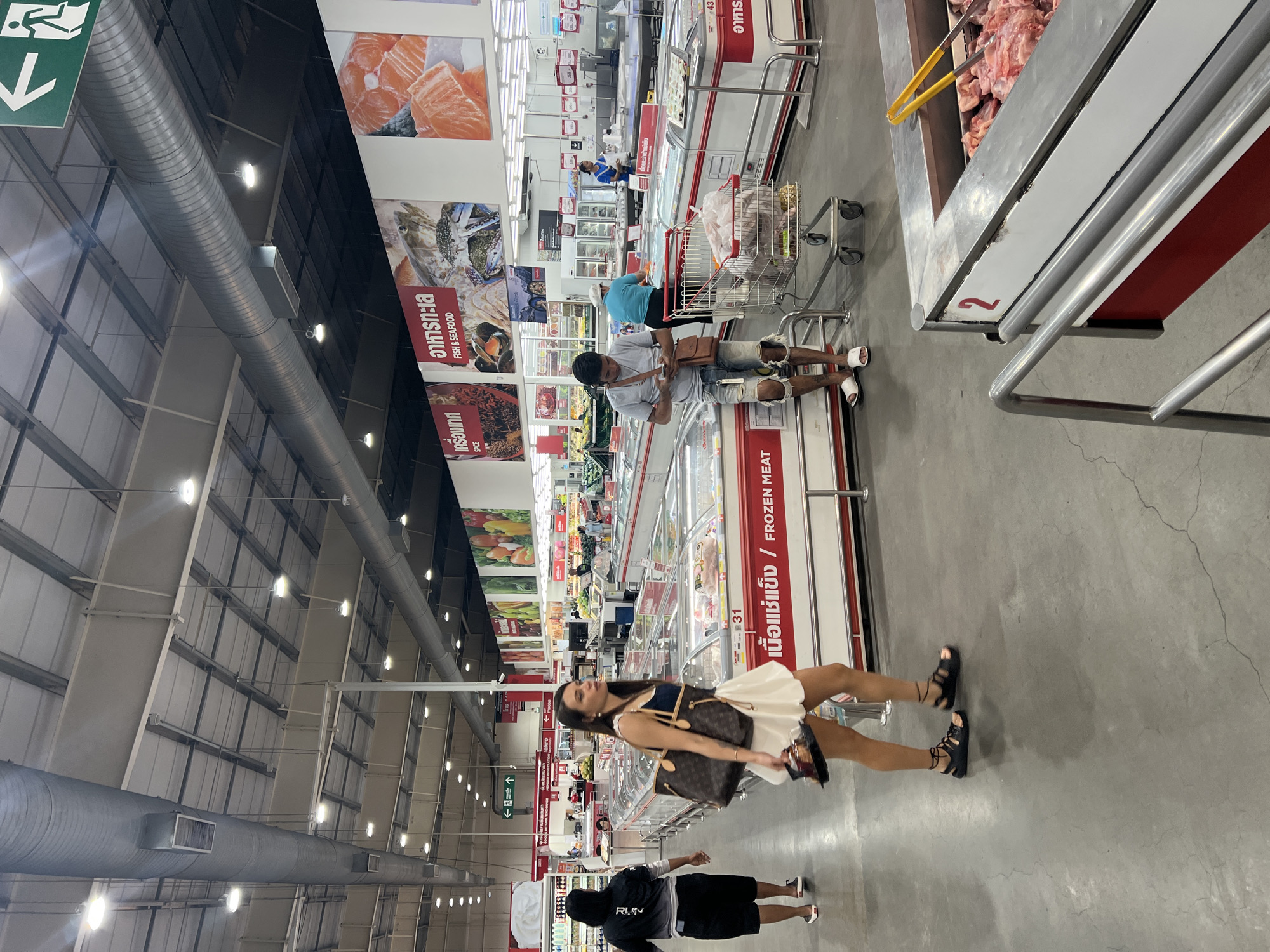 The image shows the interior of a large supermarket, likely in Baan Hua Thanon, Thailand, given the presence of Thai language signage. The ceiling features a complex array of metal beams, pipes, and lighting fixtures. Along the walls are signs with images of food items, including seafood and meat, accompanied by Thai script and English translations.
People are present in the scene, including a woman in a white dress and dark sandals, holding a brown handbag. Another person, wearing a black hoodie with the word "RUN" on the back and dark shorts, walks nearby. Further in the background, other shoppers are visible, some with shopping carts.
The scene is focused on the aisles of frozen food, marked by red and white freezers. The signs "FROZEN MEAT" and "FISH & SEAFOOD" are visible. The number "31" is prominently displayed on the side of one of the freezers, and the number "2" is seen on a meat counter.
