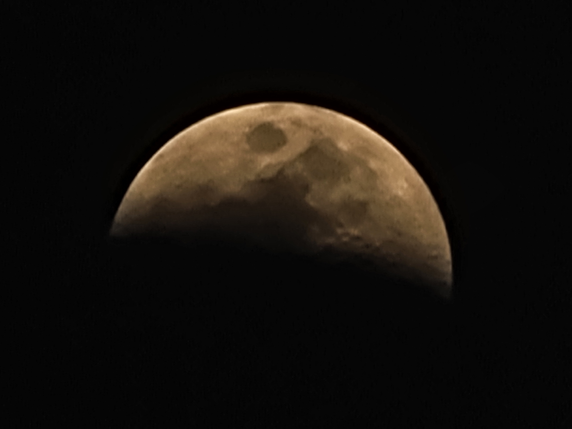 The image shows a close-up view of the moon against a black sky. Approximately half of the moon is illuminated, displaying a yellowish-brown hue. The surface of the moon reveals craters and textured features. The dark background suggests the image was taken at night. There is no discernible city or urban environment, and therefore, no specific location in Quarteira, Portugal, can be identified. The image contains only the moon and the night sky.