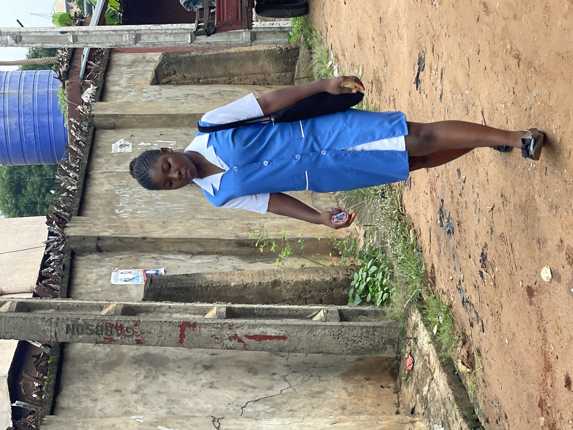 A young woman in a blue and white uniform walks on a dirt path alongside a concrete wall structure in Nkpor, Nigeria. She carries a black bag over her shoulder and holds a crumpled item in her hand. The surrounding environment includes buildings with metal roofing, overgrown vegetation, and a blue water tank. The wall bears graffiti and posters.