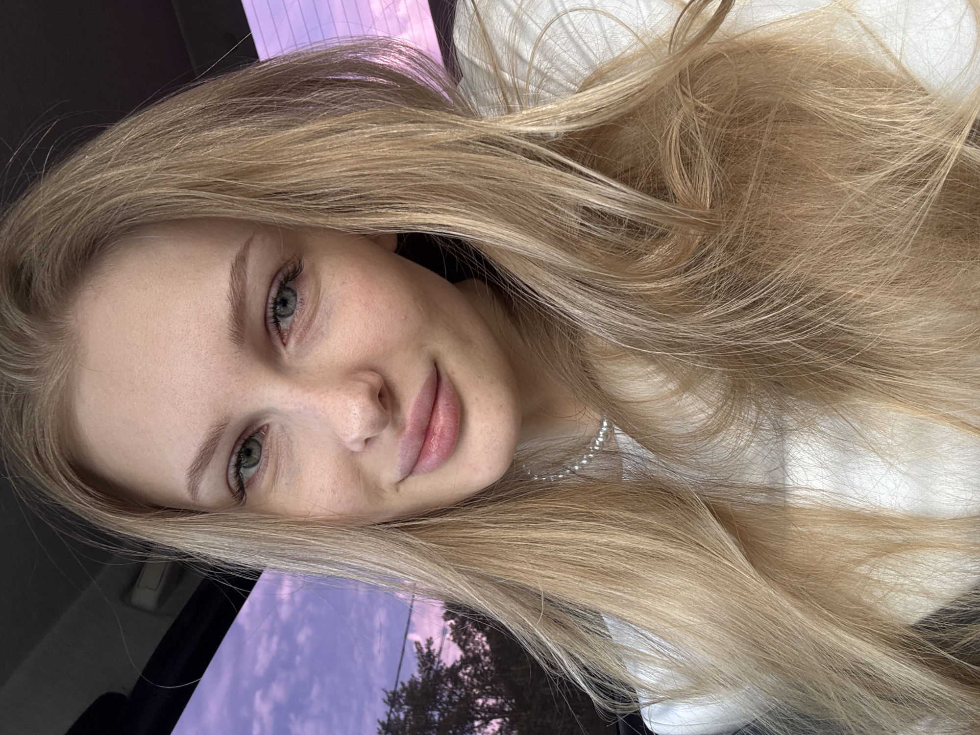 The image presents a close-up, horizontally oriented portrait of a young woman, likely a selfie. She has long, straight, light blonde hair that cascades around her face and across the right side of the frame. Her fair complexion is noticeable, with subtle freckles visible on her nose and cheeks. Her eyes are a striking green, and she has naturally full, pink lips, curving into a soft, gentle smile. She appears to be wearing a white top, complemented by a delicate pearl necklace around her neck.
The woman's head is tilted slightly, and she looks directly into the camera with a calm and serene expression. The setting suggests she is inside a vehicle, with a dark interior visible on the left side of the frame. The dominant background element is a large window reflecting an outdoor scene. The sky in the reflection displays vivid hues of purple and pink, indicating either sunset or sunrise. Silhouettes of trees are visible against this colorful sky. There is no visible text within the image. The context provided indicates the location is Perm, Russia.