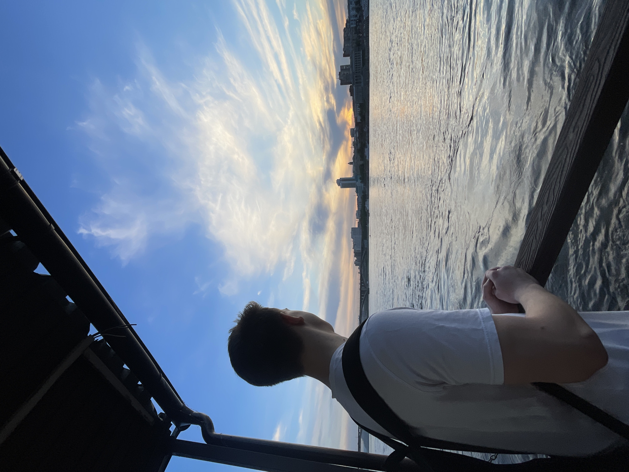 The image depicts a person from a rear-side perspective, standing by a body of water and gazing upwards and outwards towards a sprawling sky and distant cityscape. The scene is outdoors, likely during late afternoon or early evening, suggesting the golden hour or sunset.
The main subject is a person with short, dark hair, wearing a plain white t-shirt and a dark shoulder strap, possibly from a bag. They are positioned with their back mostly to the viewer, looking intently towards the horizon and sky. Their hands are clasped together and rest on a dark wooden railing that runs along the right side of the frame.
The sky is a prominent feature, transitioning from a clear, deep blue on the left to lighter blues, whites, and warm golden-orange tones towards the right horizon. Wispy, elongated clouds are streaked across the expanse, catching the warm light. Below the horizon, a distant city skyline is visible, appearing as dark silhouettes against the brighter sky and water.
A wide body of water, likely a river, fills the middle ground and right side of the image. Its surface is gently rippled, reflecting the light and colors of the sky. In the immediate foreground, a dark structure, possibly part of a boat's canopy or a viewing platform, is visible on the left, with additional dark railings or supports.
The overall atmosphere is one of peaceful contemplation, with the person seemingly engrossed in the vast, colorful sky and the expansive river view. Given the provided context, the location is Krasnoyarsk, Russia, likely overlooking the Yenisei River. There is no visible text in the image.