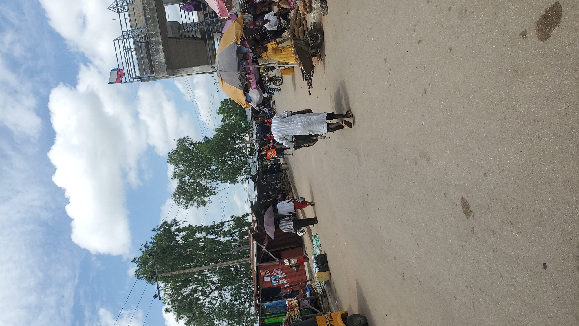 The image depicts a street market scene in Jalingo, Nigeria. In the foreground, a man in a white striped robe and dark cap pushes a wheelbarrow down a paved road. The road has several dark stains, possibly from oil or water. On the right side of the road, under a yellow and grey umbrella, a woman in a bright yellow dress and other individuals are gathered around market stalls selling produce. Several piles of root vegetables, likely yams, are visible. Further down the road on the left, a red market stall displays clothing items. Two people are walking away from the camera, one holding a dark umbrella, and a third person is walking in the same direction. In the background, several other market stalls with corrugated metal roofs line the street, with trees and utility poles visible behind them. An elevated concrete structure with railings and a flag featuring red, white, and blue horizontal stripes is visible in the upper right portion of the image, suggesting a public building or overlook. The sky is partly cloudy with scattered white clouds against a blue background. A yellow three-wheeled vehicle is partially visible on the far left.