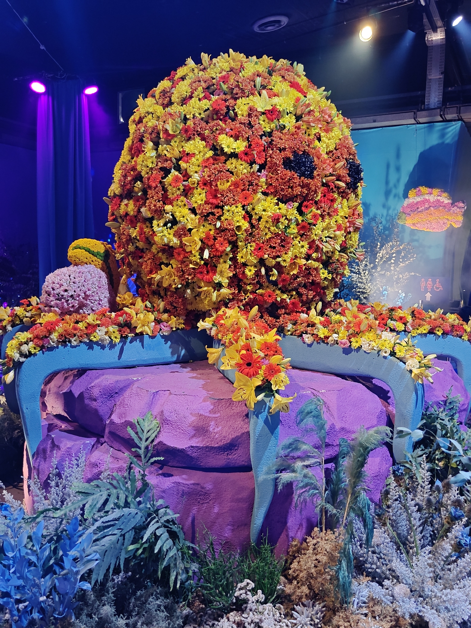 The image depicts an elaborate floral arrangement resembling a large octopus, likely a decorative display at the Tel Aviv Port (Namal Tel Aviv), a popular entertainment and shopping complex. The octopus's body is a massive sphere covered in a dense arrangement of colorful flowers, predominantly in shades of yellow, orange, and red, with some dark accents that could be dark purple or black flowers. Its tentacles appear to spread outwards, draped over a stylized, purple, rock-like structure that is accented with bright blue, curved elements. The base of the display is surrounded by various artificial plants and foliage in shades of blue and green, creating an underwater or garden-like atmosphere. In the background, there are theatrical lights, including purple spotlights, and a blue backdrop with an image that suggests water and possibly a floral depiction of a fish. The overall impression is one of a festive and artistic installation.