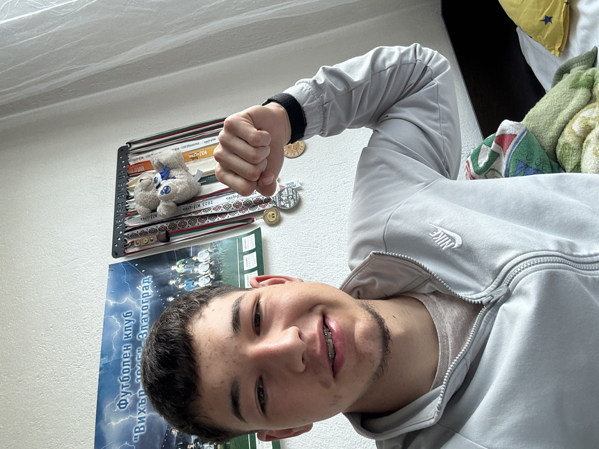A young man with braces smiles into the camera, raising his fist in the air. He is wearing a light gray Nike zip-up jacket. Behind him on the wall is a poster for "Вихър 1045" Златоград, a soccer club in Zlatograd, Bulgaria. There's a shelf above the poster with medals and small stuffed animals. The room appears to be a bedroom. The perspective suggests the picture was taken at an angle from below.
