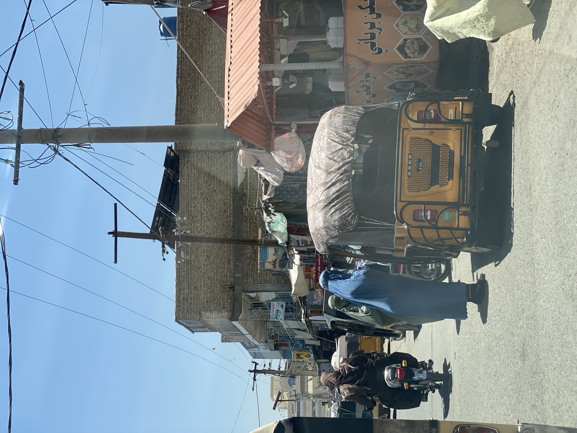 In Kandahar, Afghanistan, a street scene unfolds under a clear blue sky with power lines crisscrossing overhead. The street is paved and appears to be a commercial area, with buildings lining both sides. On the left, a woman wearing a dark hijab and brown abaya rides a red Honda motorcycle, her face obscured. Beside her, a light green auto-rickshaw is stopped. To the right of the auto-rickshaw, a woman wearing a blue burqa, covering her entire face except for her eyes, stands in the street. Behind her, another auto-rickshaw, yellow and covered with a patterned tarp, is parked.
The buildings show a mix of materials, with some constructed from brick and others appearing to have metal awnings. Signs in Arabic script are visible on the storefronts. One sign with orange lettering features images of food, suggesting a restaurant or food stall. Another building displays a white sign with blue text. A red-sloped awning covers a stall where items, possibly clothing, are displayed. The overall impression is of a bustling urban environment with active street life.