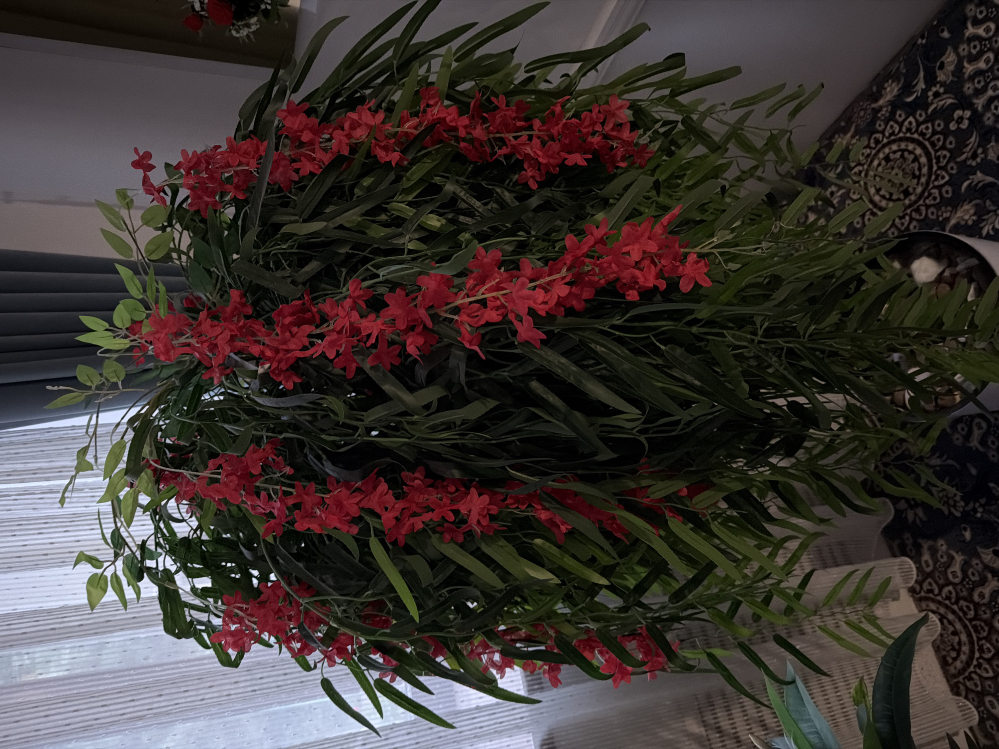 The image depicts a large, artificial hanging plant with cascading vines of small red flowers and dark green, elongated leaves. The plant is positioned in a dimly lit indoor setting, with a sheer white curtain visible in the background to the left and a dark grey curtain behind it. The lower portion of the image shows a section of a patterned rug with an intricate design in dark colors. No people are visible in the image. The scene appears to be a domestic interior. The location within the city of Khumsan, Uzbekistan is not discernible from the visual information provided.