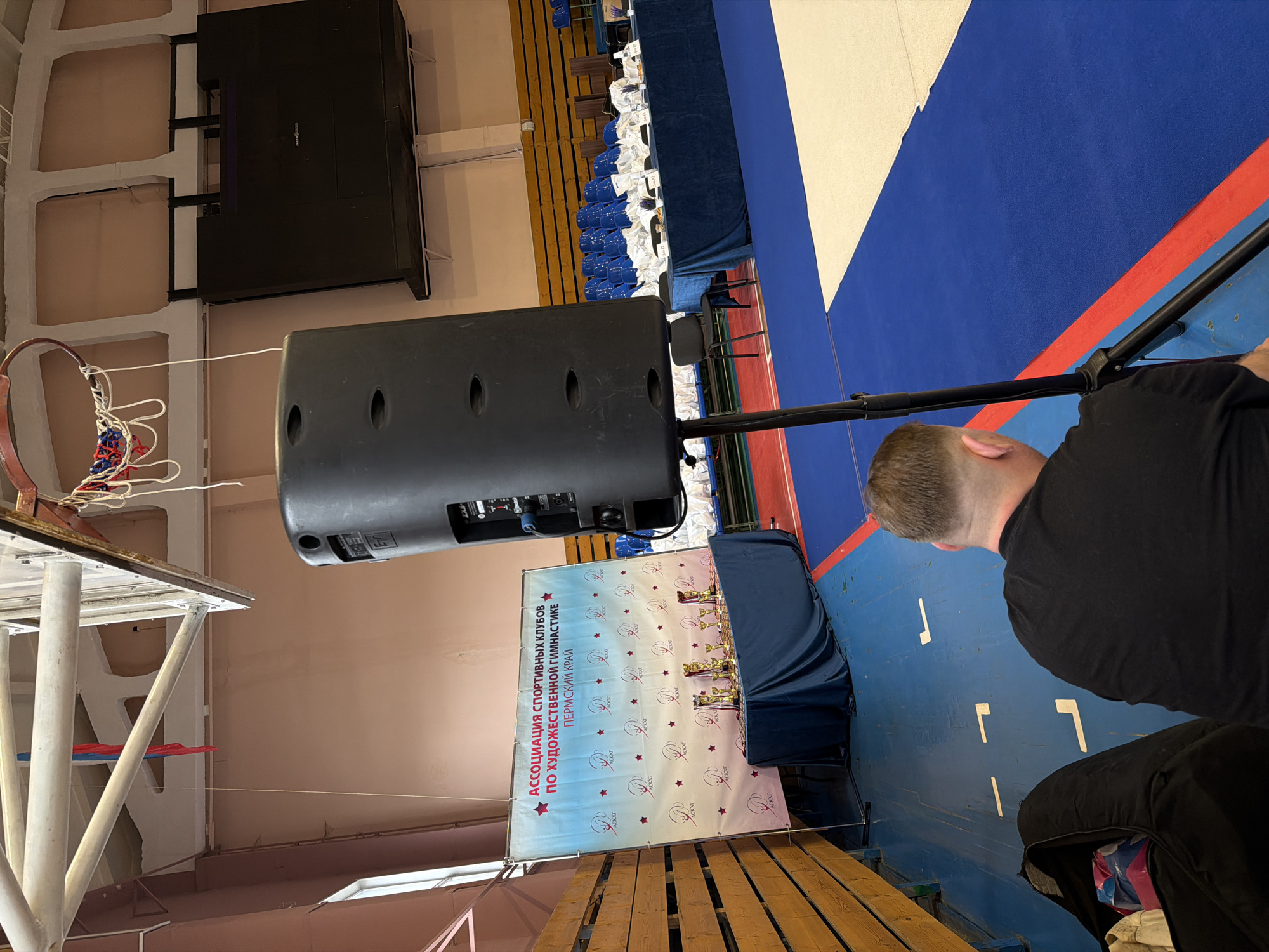 Here is a forensic description of the image:
The image shows an indoor event space, possibly a gymnasium, likely in Perm, Russia, based on the presence of the banner.
**People:** The back of a person's head and a black-clad torso are visible in the lower right.
**Subjects:** There are several objects within the gymnasium. A large black speaker is prominently placed. Behind it, there is a banner promoting an association of sports clubs for rhythmic gymnastics in the Perm region. There are also trophies visible on a table covered in blue cloth in front of the banner.
**Objects:** Besides the speaker, banner, trophies, and table, there's a basketball hoop in the background. The floor is blue with white and red markings, likely for sports activities. On the left is some wood paneling. There are rows of blue chairs. An electronic display screen hangs from the ceiling.
**Actions:** None.
**Interactions:** None explicitly visible.
**Environment:** The gymnasium appears well-lit and spacious. The overall impression is of a setup for a sports-related event, possibly a competition or awards ceremony.