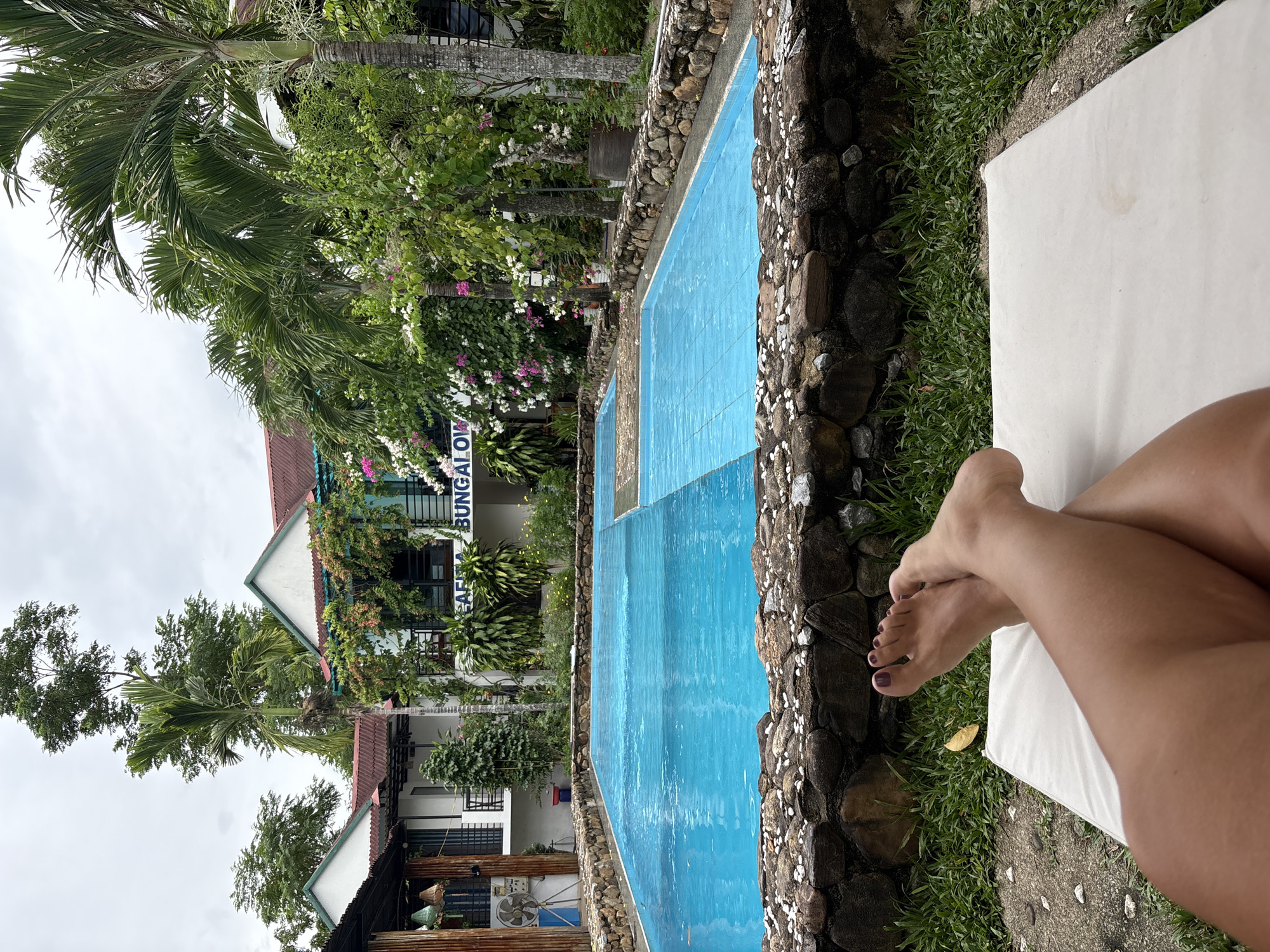 A person's legs and feet are visible in the foreground, resting on a white lounger in a tropical outdoor setting. Behind the lounger, a bright blue rectangular swimming pool with a stone border is the central focus. The pool is divided into two sections by a submerged line. Beyond the pool, a two-story building with white walls, red roofs, and balconies is visible. Lush green trees, palm trees, and flowering vines with pink and white blossoms surround the building. A sign on the building reads "CAFÉ BUNGALOW." The environment appears to be a resort or guesthouse in Bố Trạch Commune, Vietnam. The sky is overcast, suggesting a cloudy day.