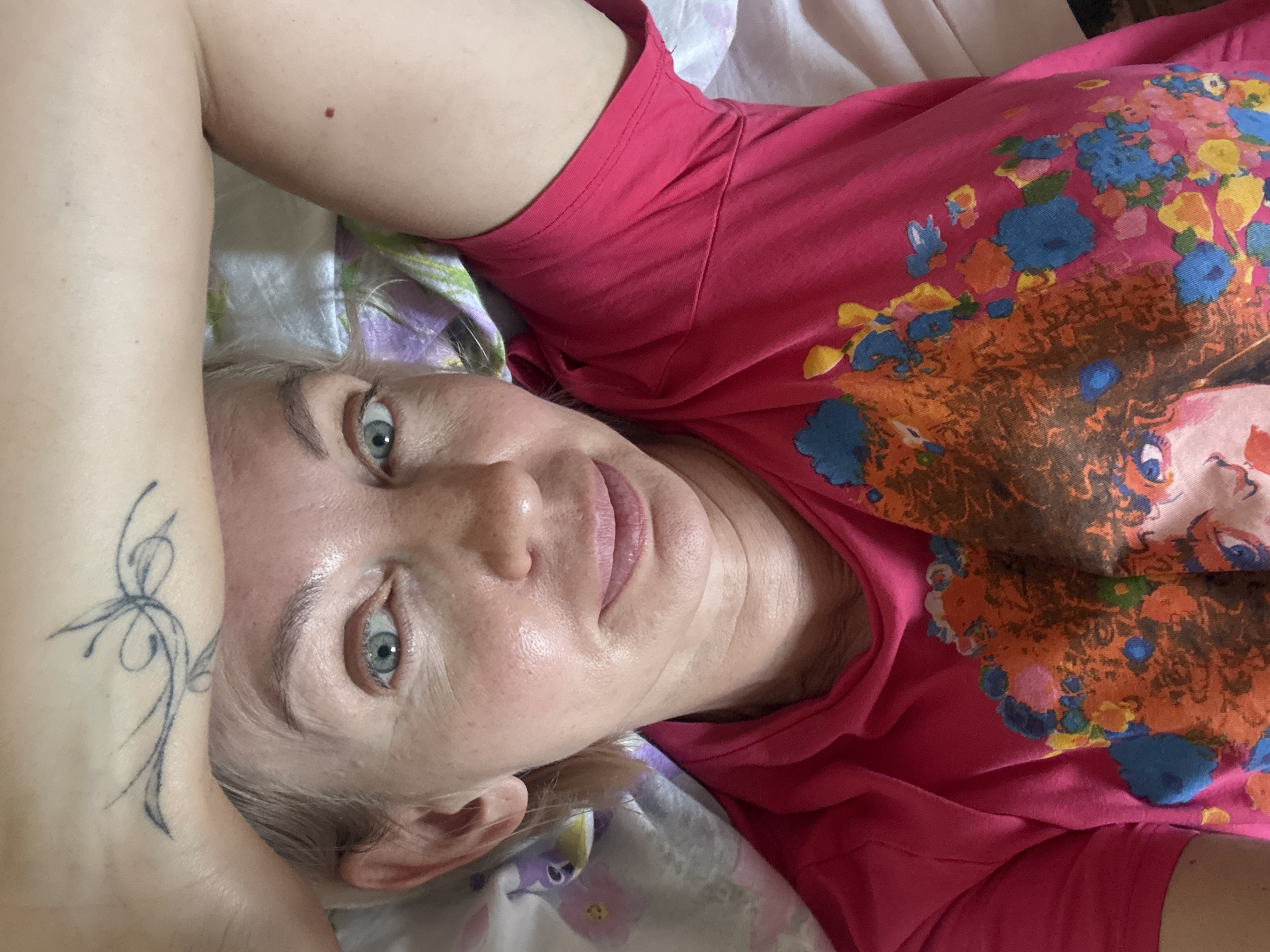 A woman with blonde hair and blue eyes lies in bed, holding her arm over her head. She is wearing a bright pink t-shirt with a colorful abstract print on it. The bed appears to be covered in floral patterned sheets. A tattoo is visible on her forearm. A small red mole is visible on her upper arm. The scene is an indoor setting, possibly a bedroom. The location is not specified to be in Sochi, Russia.