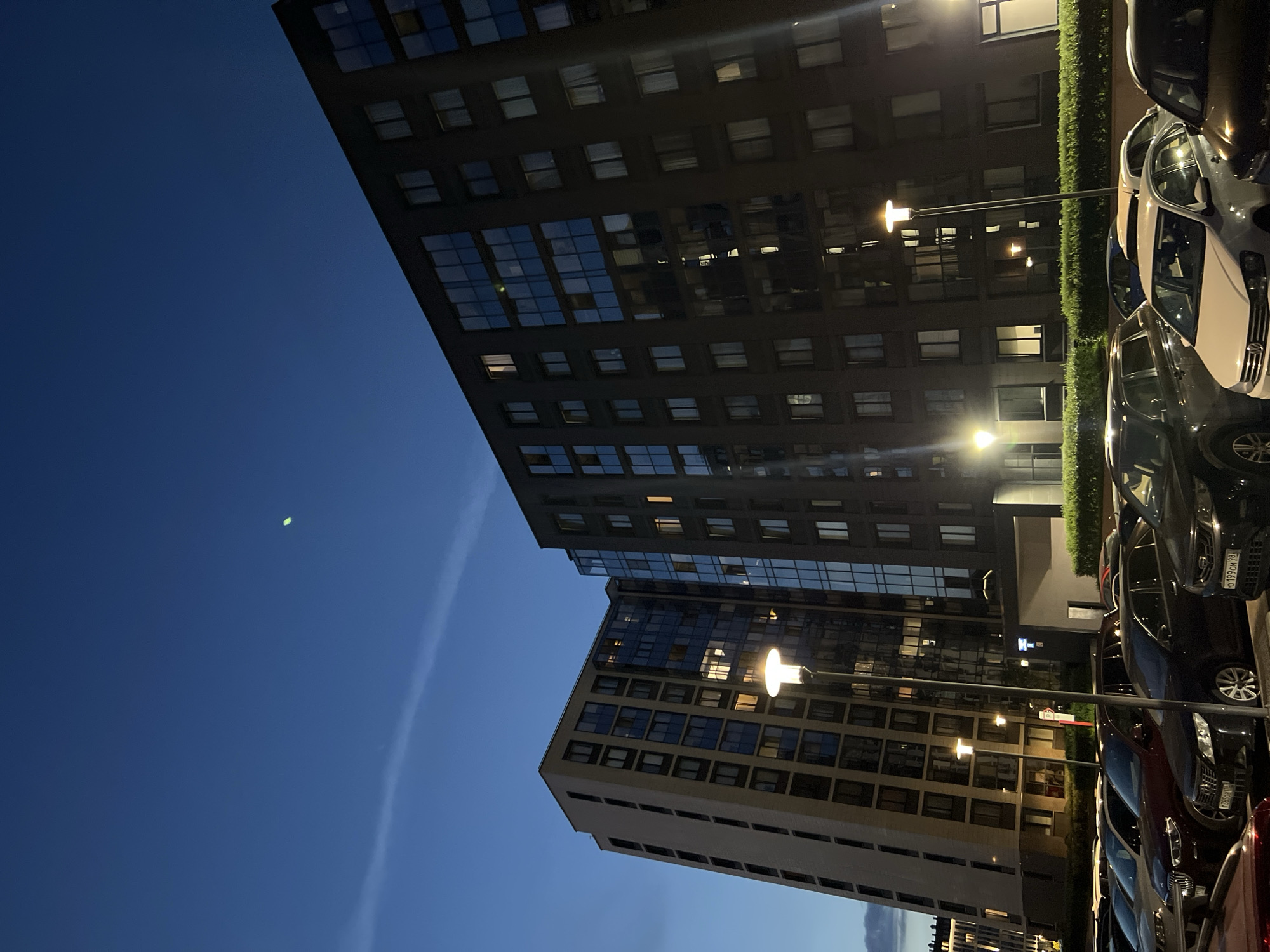 This is a low-angle, outdoor, full shot of modern apartment buildings at dusk. The sky is a deep blue with a thin, wispy cloud formation and a faint green light streak. Two tall buildings are visible; one is light-colored with many windows, and the other is dark with reflective glass windows. Several floors of windows are illuminated from within, suggesting occupancy. A modern street lamp with a lit top stands in the foreground. Below the buildings is a parking lot filled with various cars. The environment appears to be an urban or suburban residential area. There are no people visible in the image. The overall atmosphere is serene and slightly futuristic.