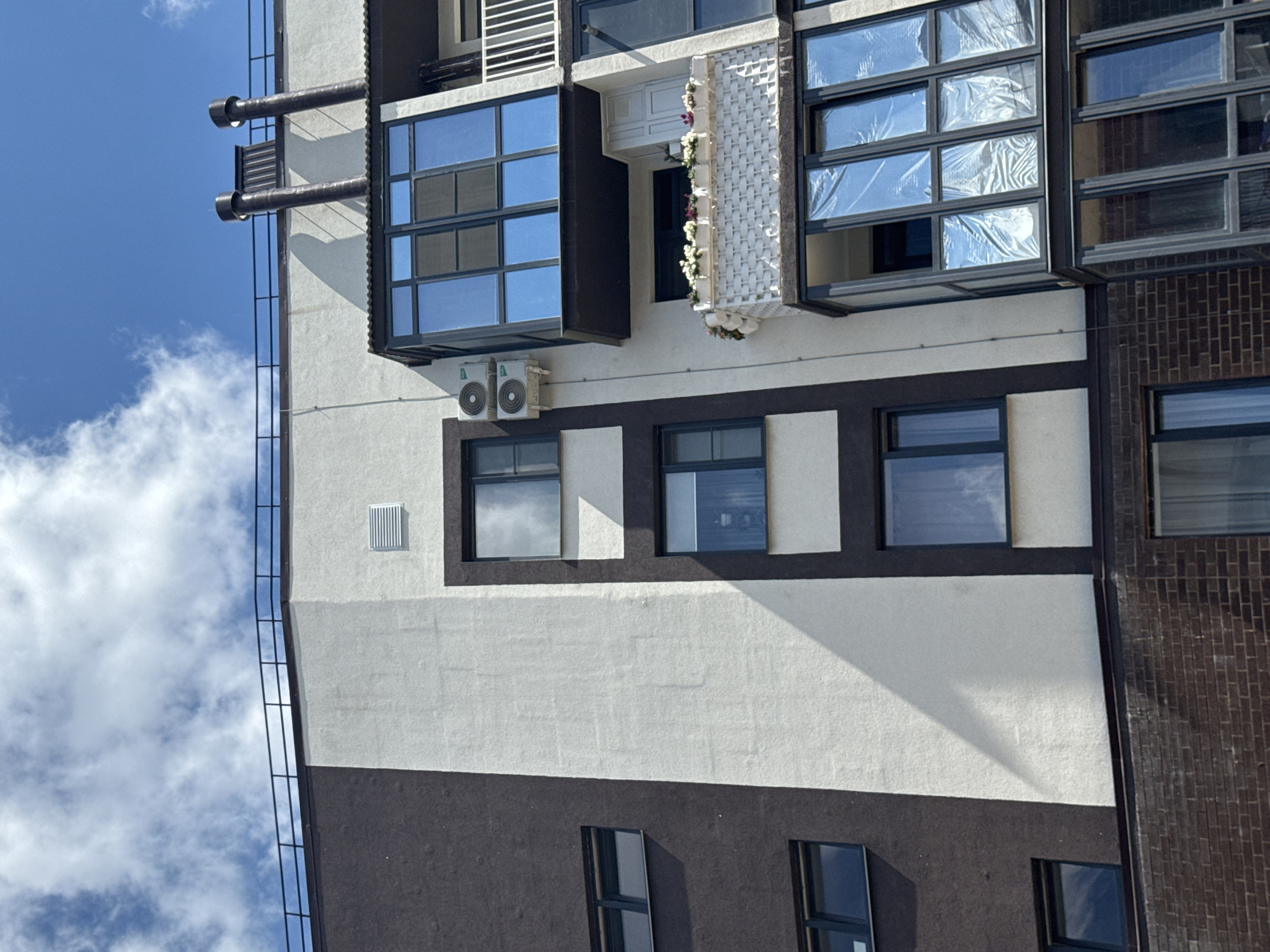 The image shows the facade of a modern multi-story building against a bright blue sky with scattered white clouds. The building's exterior features a distinct pattern of dark brown and off-white sections. Several windows of varying sizes are visible, some with dark frames and reflective glass, others with lighter frames and tinted glass.
On the right side of the building, a distinctive feature is a multi-level balcony or sunroom with large, floor-to-ceiling windows. The upper section of this feature is enclosed and appears to have blue-tinted glass panels. Below it, a smaller balcony is adorned with numerous white planters filled with flowers. A white railing is also visible on the right side of the building, extending upwards.
Two air conditioning units are mounted on the off-white section of the building, positioned side-by-side. The roofline is topped with a dark railing, and two dark, cylindrical ventilation pipes or chimneys protrude from the roof on the right side. The lower portion of the building visible in the image is constructed of dark brown brickwork.
The scene is devoid of any people or visible activity. The lighting suggests it is daytime, likely sunny, with a strong shadow cast diagonally across the off-white section of the building from an unseen structure or architectural element. The architectural style and the presence of multiple windows and balconies suggest a residential apartment building. The image is taken from a low-angle perspective, looking upwards.