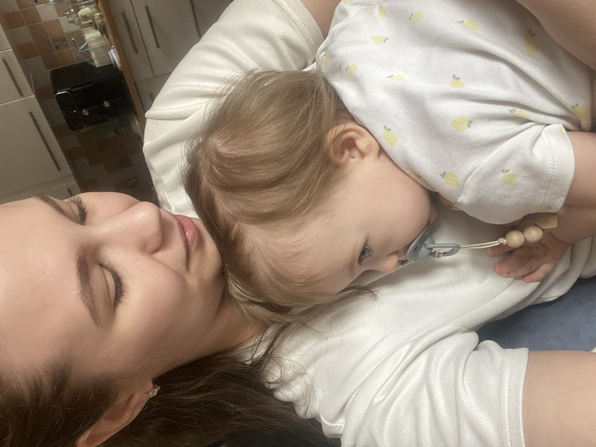 This image depicts an adult female holding an infant in an indoor residential setting, specifically a kitchen, located in the city of Chaikovsky, Russia. The adult female, positioned on the left side of the frame, has light brown hair, wears a light-colored short-sleeved top, and has her head tilted downward, looking towards the infant. A small earring is visible in her right ear. The infant, with light brown hair, is clad in a white onesie adorned with yellow lemon patterns, and is held against the adult's left side, resting its head on the adult's chest/shoulder area. A blue pacifier, attached to a clip featuring wooden beads, is present in the infant's mouth. The background reveals light-colored kitchen cabinetry with horizontal silver handles, a tiled backsplash, and a dark-colored kitchen appliance on a countertop.
