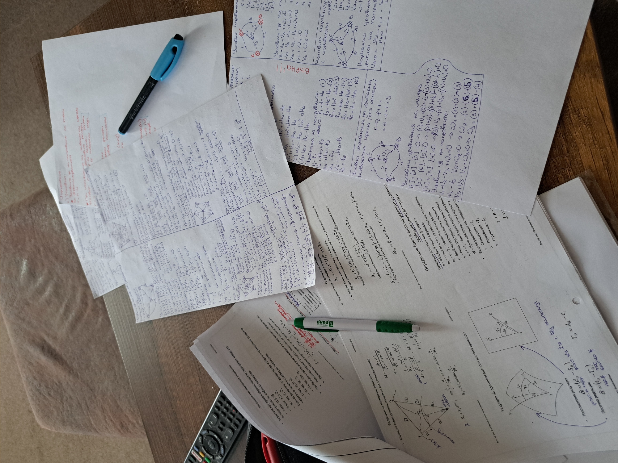The image shows a table cluttered with papers filled with handwritten notes and equations in both English and another language, possibly Bulgarian based on the Cyrillic script. A blue and black pen lies on one stack of papers, while a green and white pen rests on another. A television remote control is visible in the bottom left corner. The scene appears to be a study or workspace, possibly in a home, though the specific location is unknown.