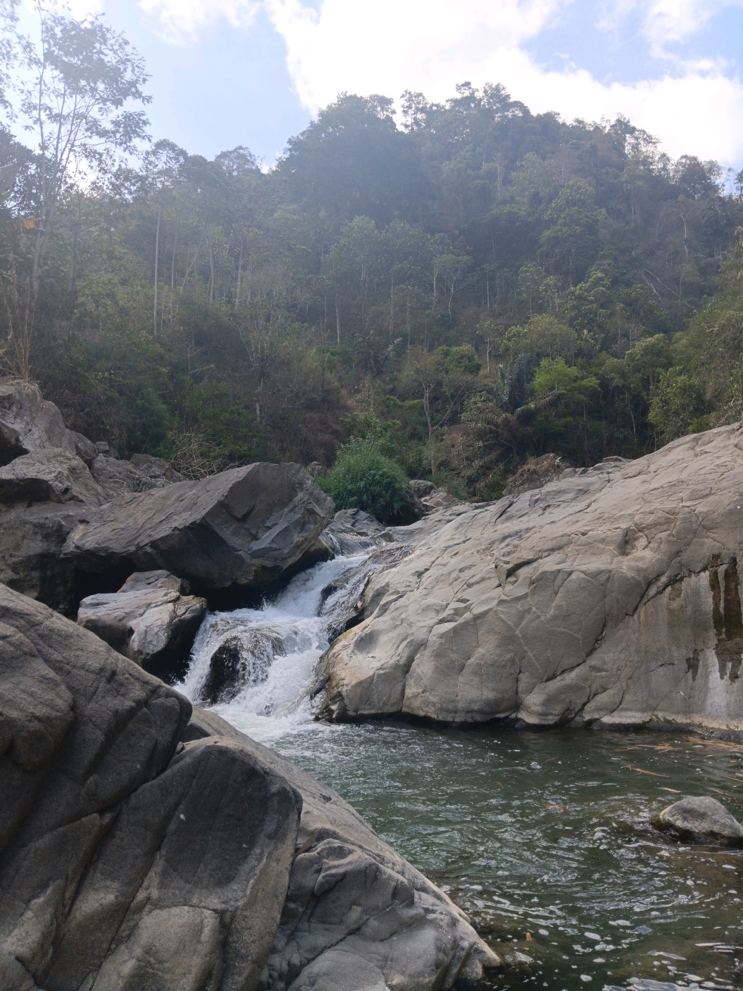 The image depicts a natural scene of a rocky waterfall in a forested mountain area. The foreground is dominated by large, weathered grey boulders, some of which are partially submerged in the dark, rippling water of the stream. A small waterfall cascades over rocks in the middle ground, creating white foam and spray. The background features a dense, green forest covering a steep mountainside, with trees of varying heights and foliage. The sky above is partly cloudy, with patches of bright white clouds against a pale blue backdrop. The overall impression is one of a secluded, natural environment. The image does not contain any people, specific buildings, or identifiable man-made structures, suggesting it was taken in a wilderness area, likely within the natural landscapes surrounding Sinduaji, Indonesia.
