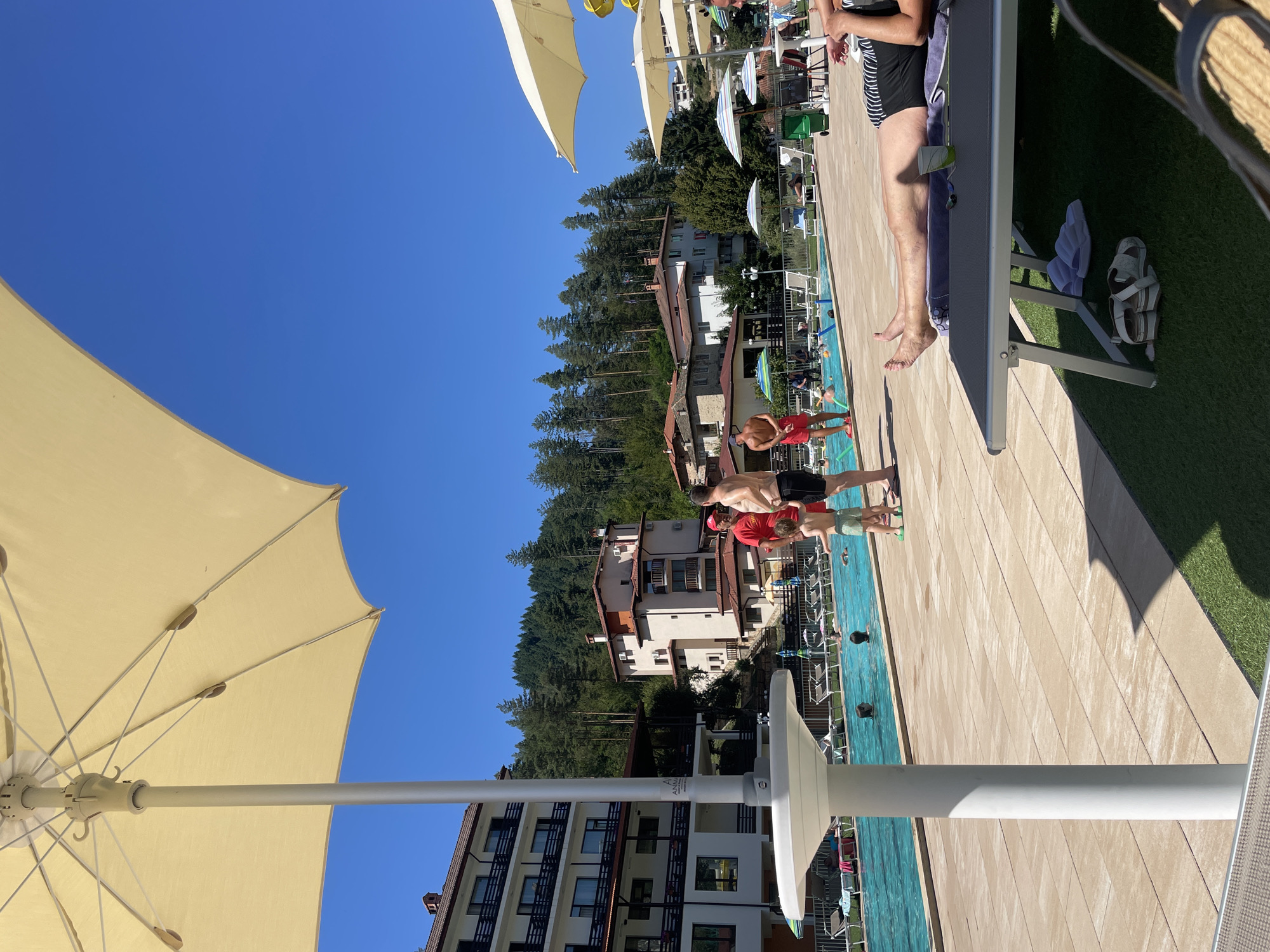 The image depicts a sunny outdoor scene at a resort in Banite, Bulgaria, likely a hotel with a swimming pool. In the foreground, a large, light-colored umbrella shades a paved area. To the left, a multi-story hotel building with white facades and dark balconies is visible. Beyond the paved area is a bright blue swimming pool, with several people swimming and wading. On the poolside, lounge chairs are arranged, some occupied. In the midground, several other buildings with red-tiled roofs are situated amongst trees. A group of people, including a man in a red shirt, a man in black shorts, and a young child, are standing near the edge of the pool. In the background, a dense forest covers a hillside. The sky is clear and blue. The overall impression is of a leisure resort on a bright, warm day.