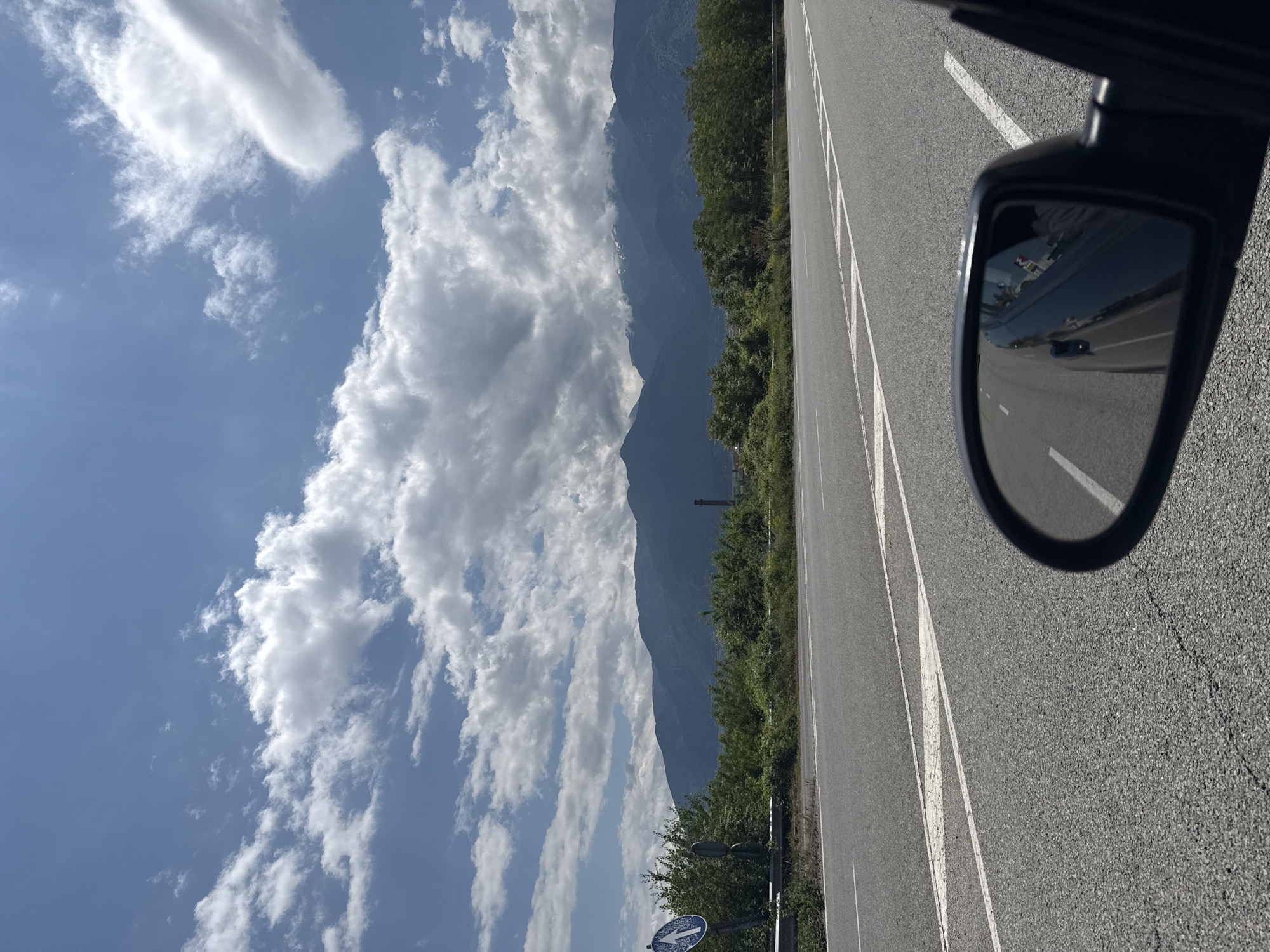 The image captures a scenic view from inside a moving vehicle, looking towards the left side of the road. In the foreground, the asphalt road stretches diagonally, featuring white painted lines, including a solid line on the shoulder and dashed lines marking lanes. The right side mirror of the car is prominently visible in the lower right, reflecting a distorted view of the car's interior (possibly a dashboard or passenger) and the road receding behind.
The midground features lush green trees and bushes bordering the road on the left. A partially visible round blue road sign with a white arrow pointing upwards (or slightly left, depending on the exact orientation) is visible amidst the vegetation. Beyond the trees, a majestic range of dark, imposing mountains dominates the background, their peaks stretching across the horizon. Above the mountains and extending over the scene is a vibrant blue sky dotted with scattered white, fluffy cumulus clouds, suggesting clear and pleasant weather.
The scene depicts bright daylight, likely late morning or early afternoon, given the intensity of the light and the clear skies. The overall impression is that of a journey through a picturesque, mountainous landscape. No discernible text is clearly visible on any signs or objects within the image, either in the main view or in the mirror's reflection. The context indicates the location is Piperevo, Bulgaria.