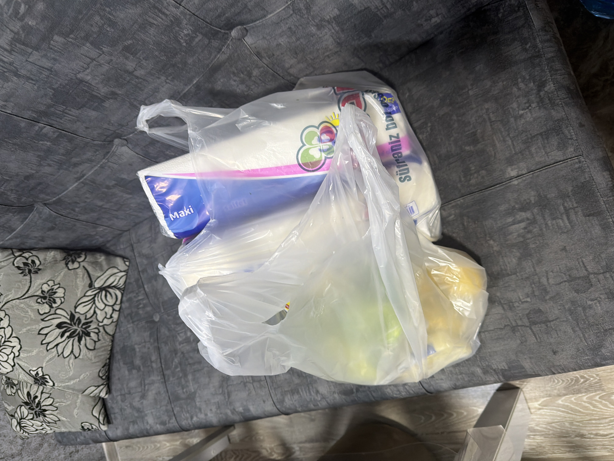A clear plastic grocery bag rests on a dark gray tufted sofa, containing a roll of paper towels and what appears to be produce. The paper towel roll is labeled "Maki" in blue and pink lettering. The bag also has visible writing in Turkish, reading "Süfreniz bol". Two decorative pillows with a floral pattern in gray and white are placed on the left side of the sofa. The lighting suggests an indoor environment. There is no indication of the city of Digah, Azerbaijan.