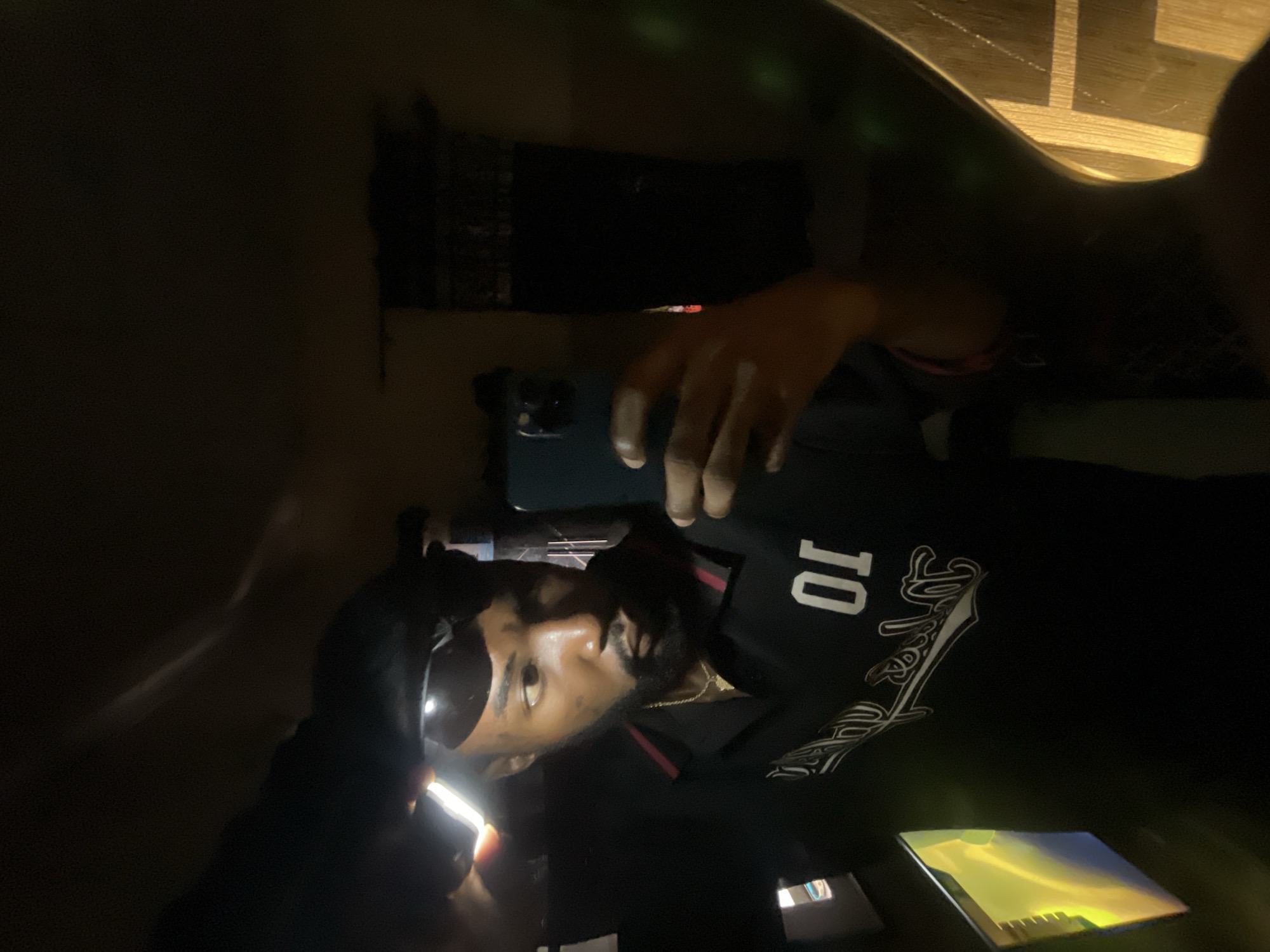 A male individual is depicted in a mirror selfie, captured indoors in a dimly lit environment, within what appears to be a private residence in Lokoja, Nigeria. The subject has dark skin, a short beard, and is wearing sunglasses positioned on his forehead. He is dressed in a black polo shirt with a red and black striped collar, featuring white numerals "01" on the right chest and a white script design below. A gold-colored chain is visible around his neck. He is holding a dark-colored smartphone in his right hand, oriented towards the mirror to record his reflection. A distinct, bright, rectangular light source is reflected in the upper left portion of the mirror, partially illuminating the subject's head. The reflected background shows a dark curtain or fabric on the right, and a visible wooden surface below it. To the lower left, a rectangular object exhibiting a yellowish-green glow is also reflected.